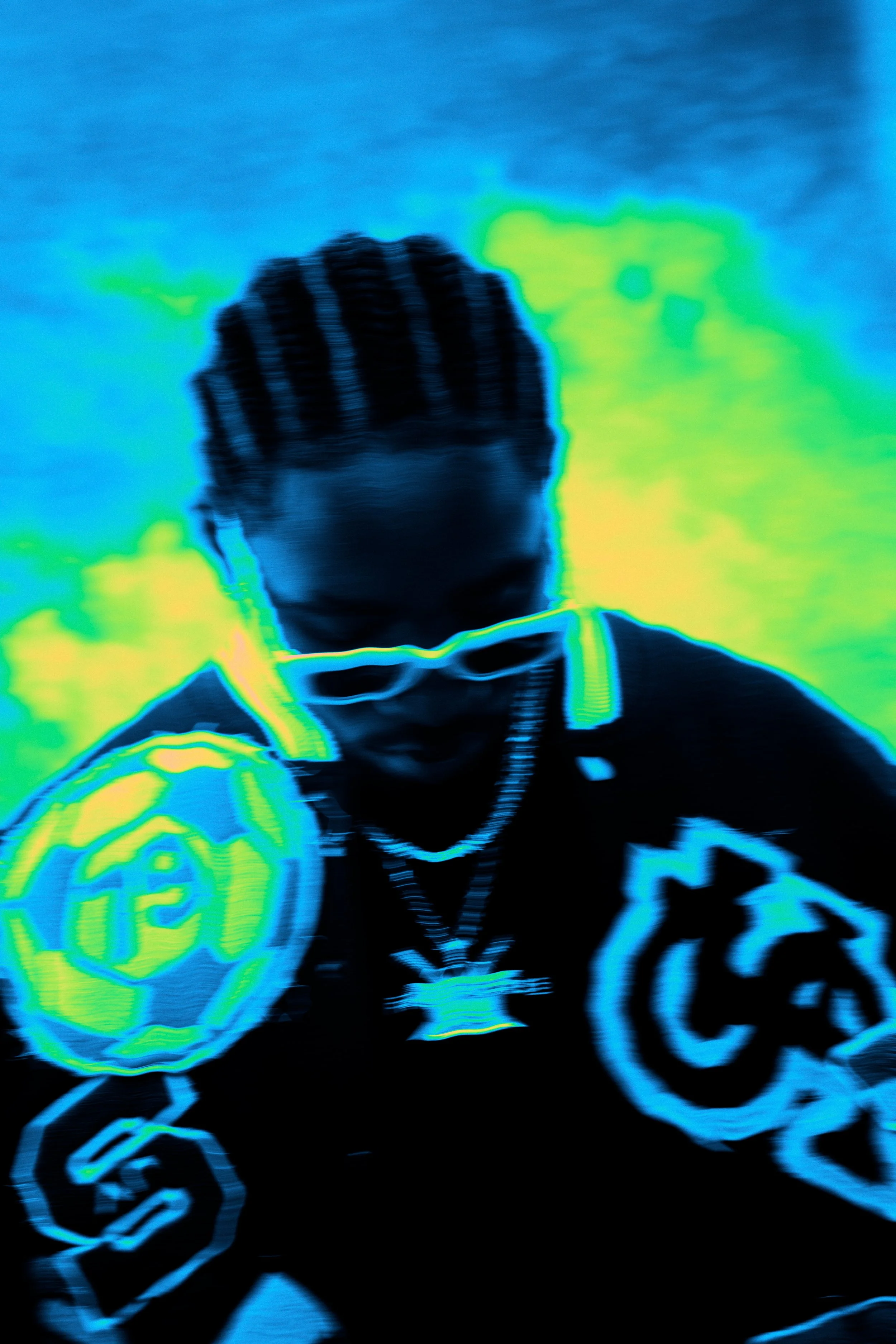 Person with braided hair wearing sunglasses, jewelry, and a jacket with logos, looking downward, UV or infrared effect applied.