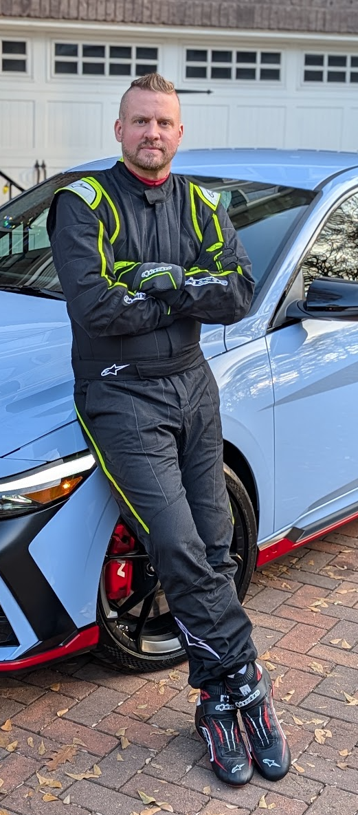 A man in racing gear leaning against a light blue sports car on a brick driveway.