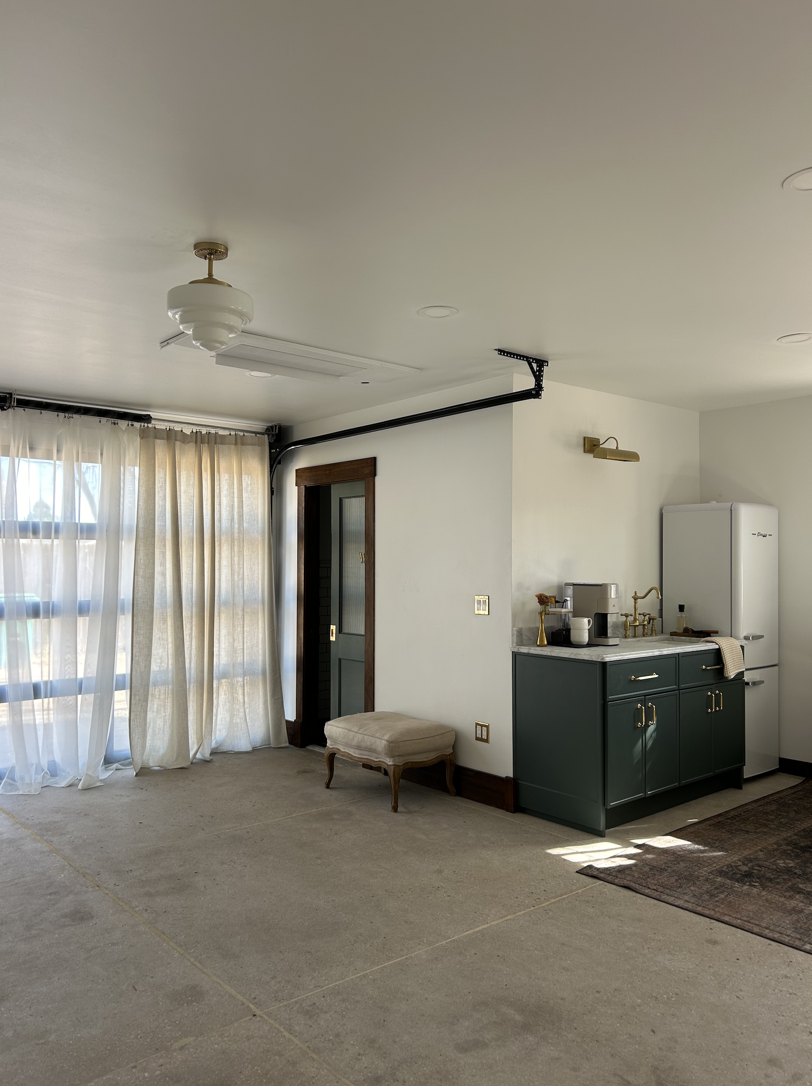 Living room with beige carpet, white curtains covering large windows, a small beige upholstered bench, green kitchen cabinet with gold handles, white refrigerator, coffee station, and a dark area rug.