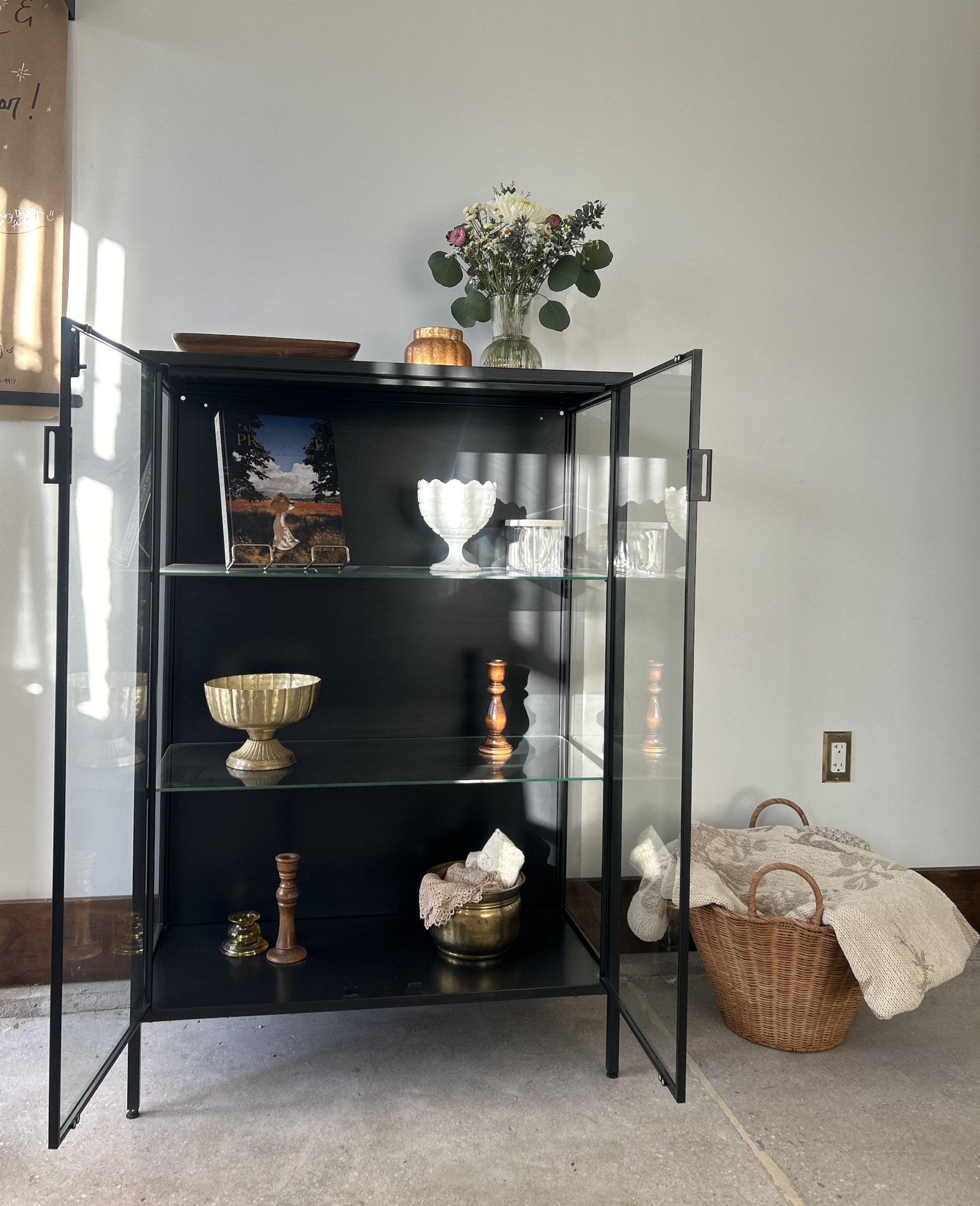 Black display cabinet with glass doors containing decorative bowls, candlesticks, and a book; on top of the cabinet is a glass vase with flowers, a small wooden bowl, and a small gold container; a wicker basket with cloth is on the floor nearby.
