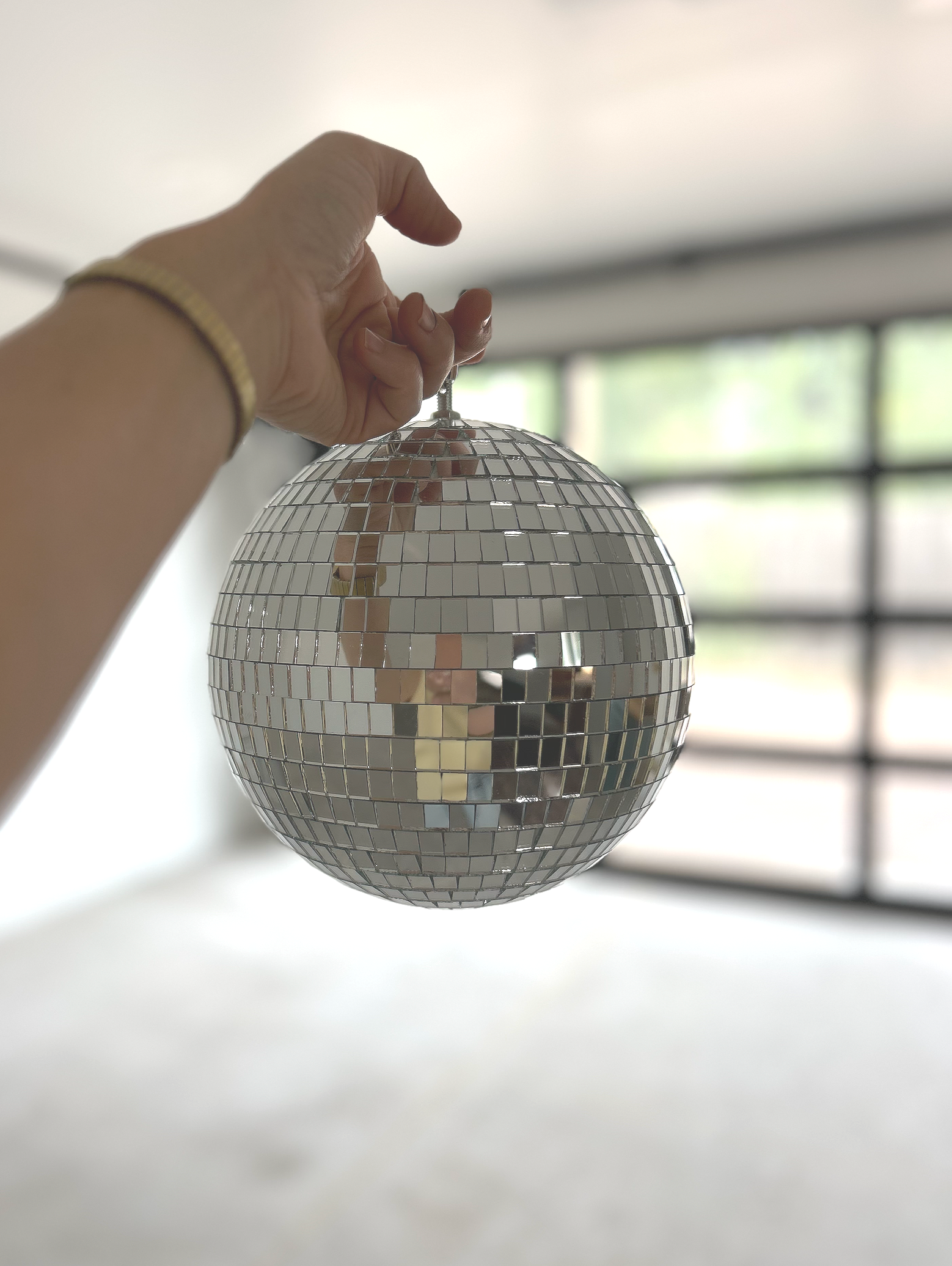 A hand holding a silver disco ball in a room with large windows and neutral-colored walls.
