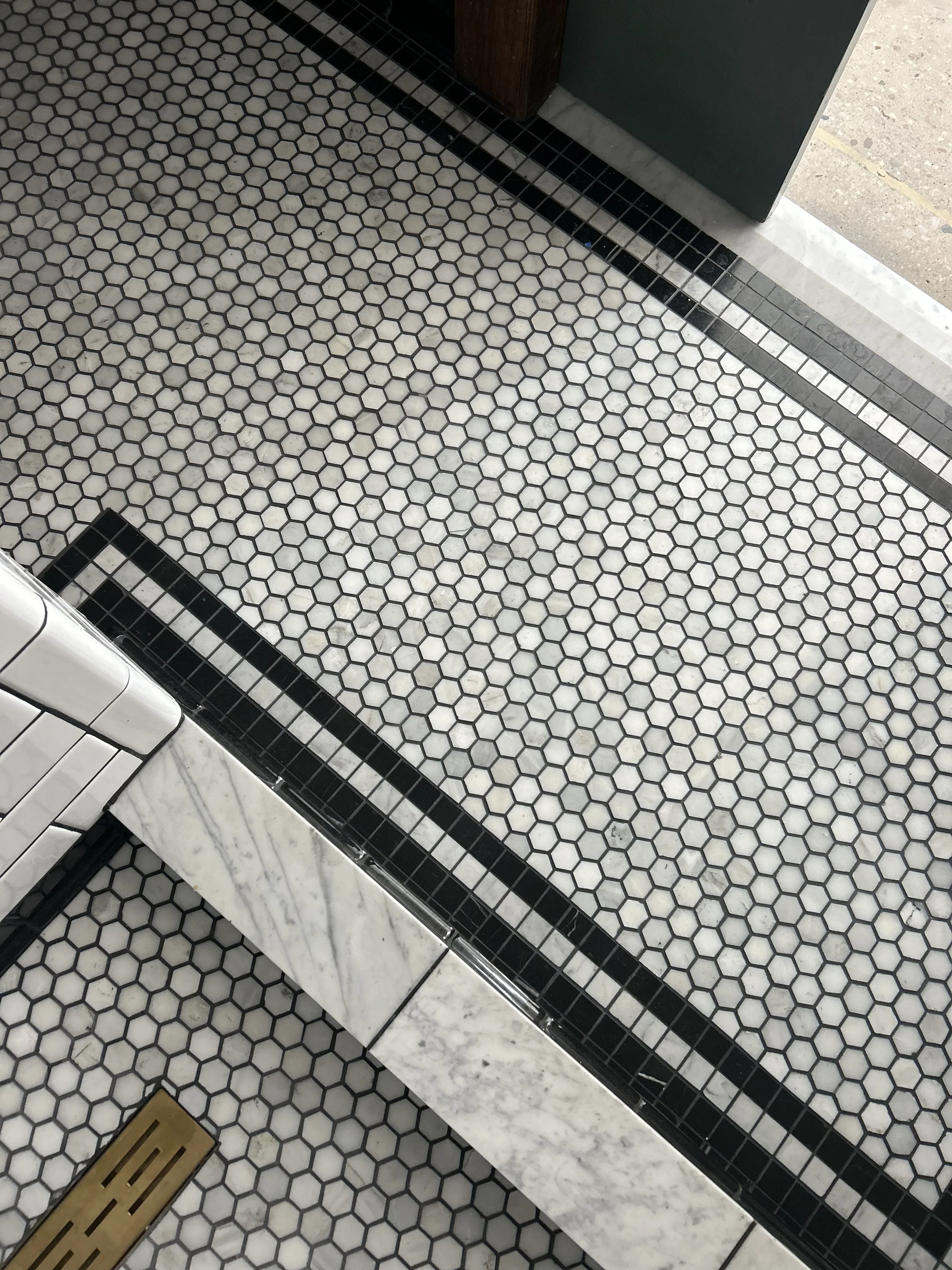 Close-up of white and black hexagonal tile floor with marble border, a black metal drain cover, and a section of a wooden bench and a green door frame.