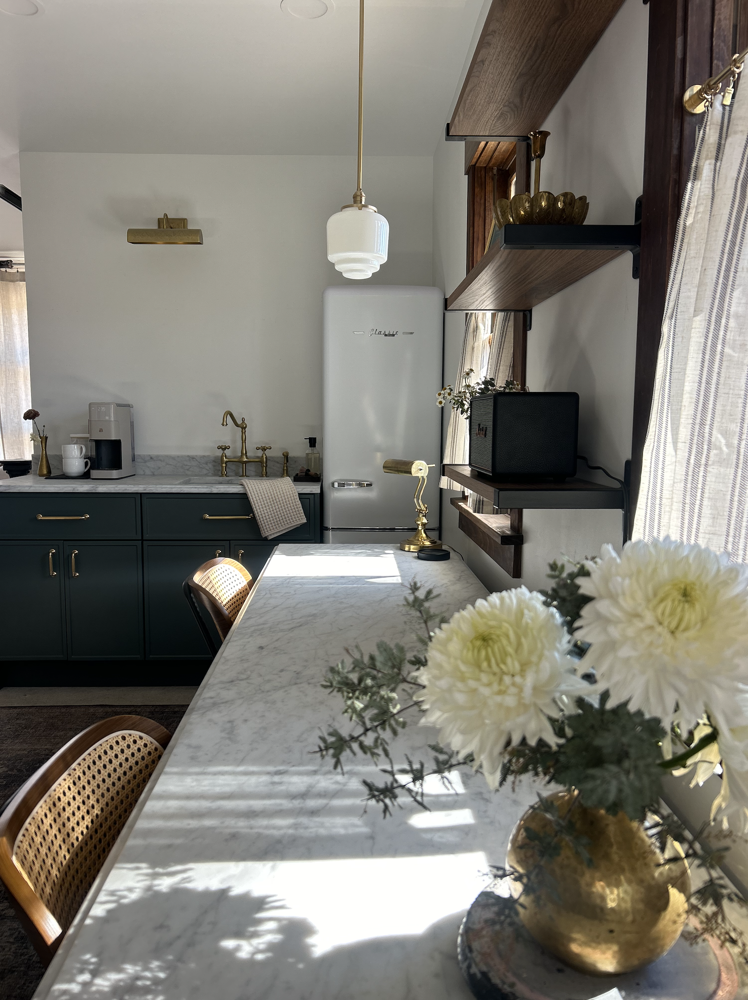 Bright, modern kitchen with a marble countertop, green cabinetry, and gold fixtures. A vase with white flowers is on the countertop near a large window with curtains. An old-fashioned refrigerator and open wooden shelves hold decorative items and a s