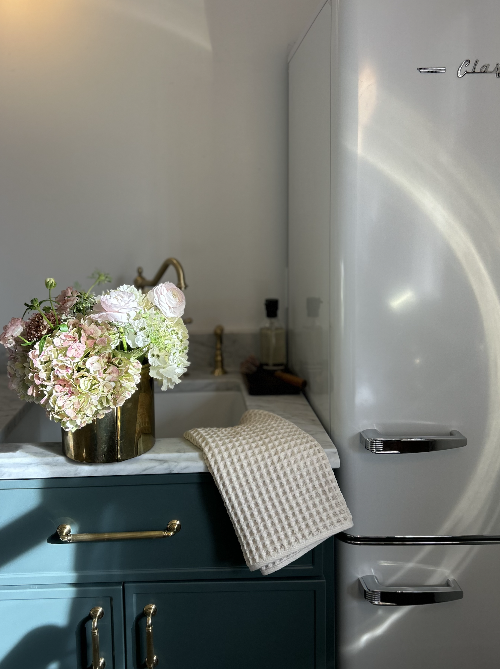 A kitchen scene with a teal cabinet, a marble countertop, a brass faucet, a bouquet of pastel flowers in a brass vase, and a beige textured towel draped over the counter. A vintage silver refrigerator is in the background.