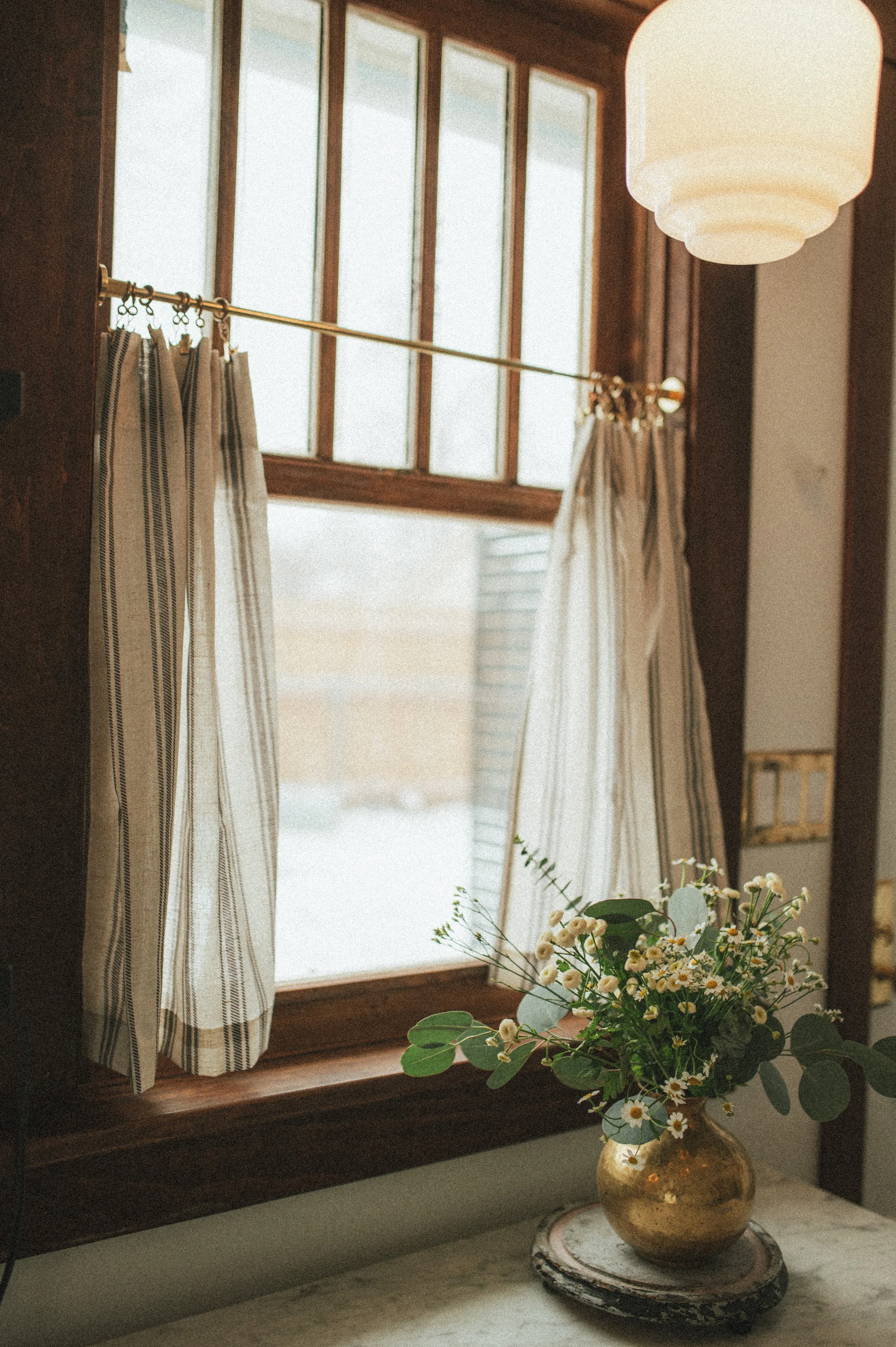 A window with wooden frames and striped curtains, a white hanging lamp, and a gold vase with white and yellow flowers on a marble surface.