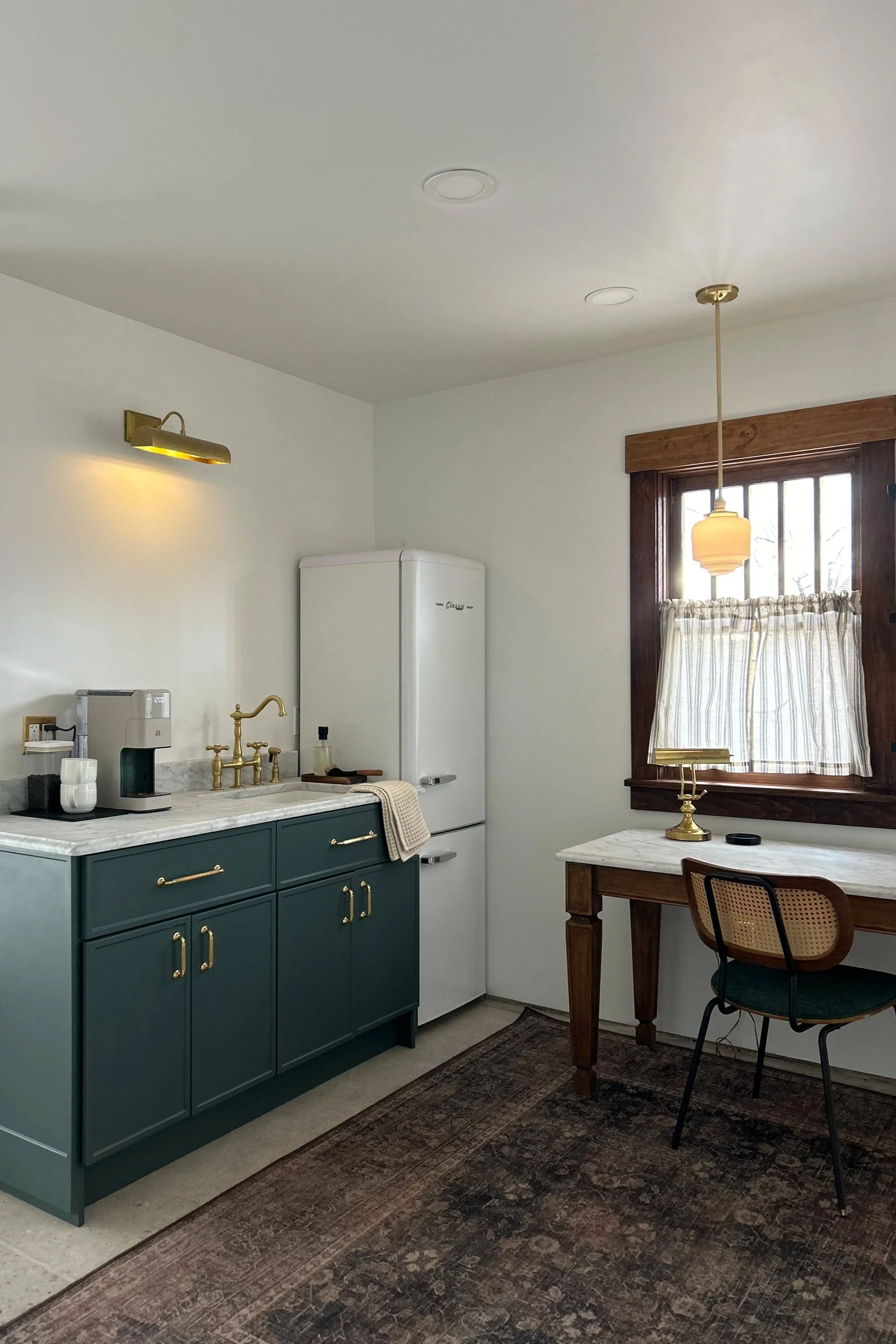 A kitchen corner with a teal cabinet, gold faucet, coffee machine, white refrigerator, wooden desk, vintage chair, window with striped curtain, and hanging lamp.