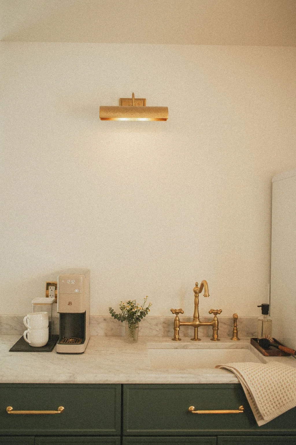 A kitchen counter with a coffee maker, cups, a small vase with flowers, a gold faucet, a sponge, and a towel, with a minimalistic beige wall and light fixtures.