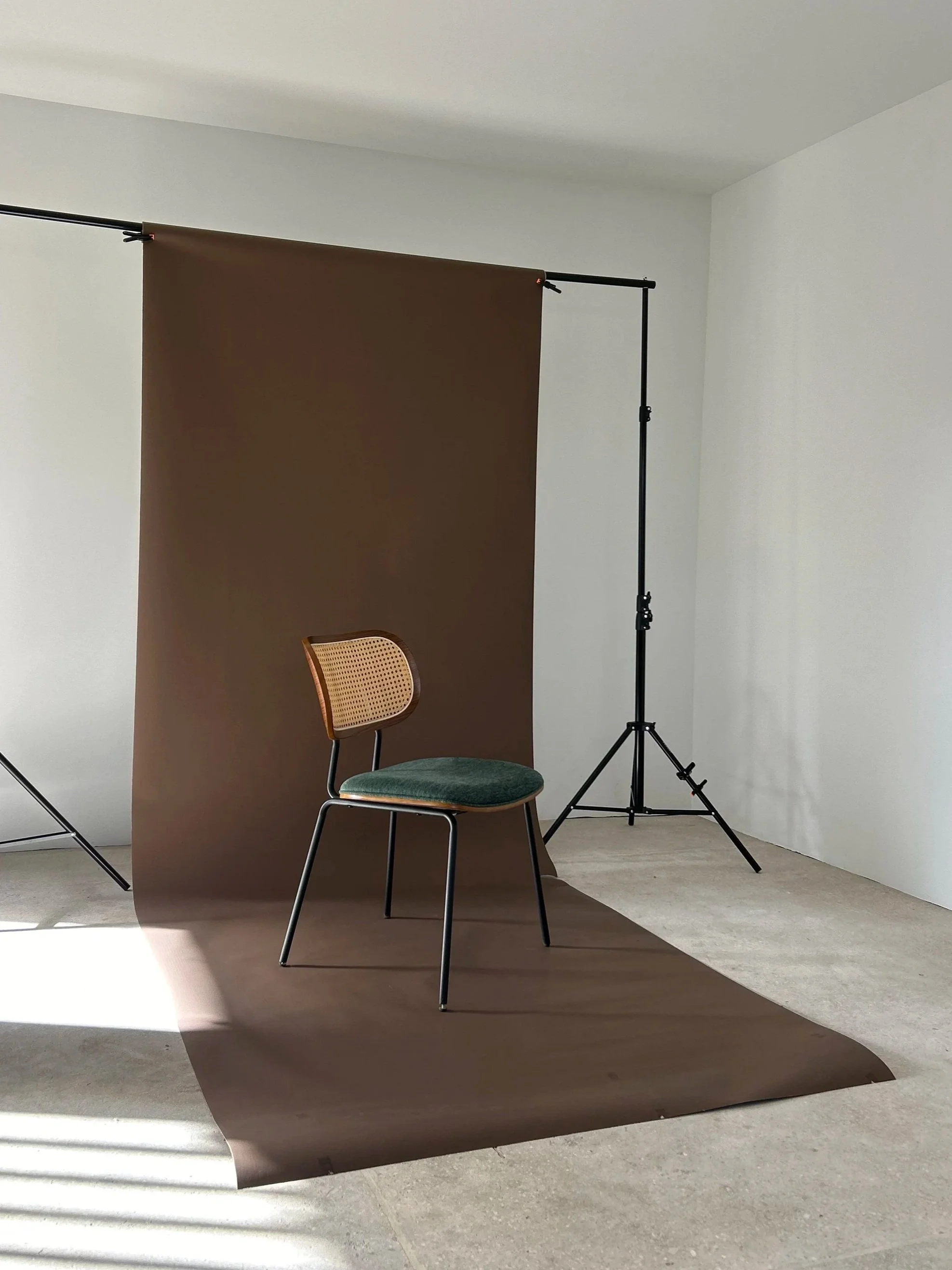 Indoor photography studio setup with a green cushioned chair in front of a brown backdrop, supported by a black stand, with natural sunlight casting shadows on the floor.