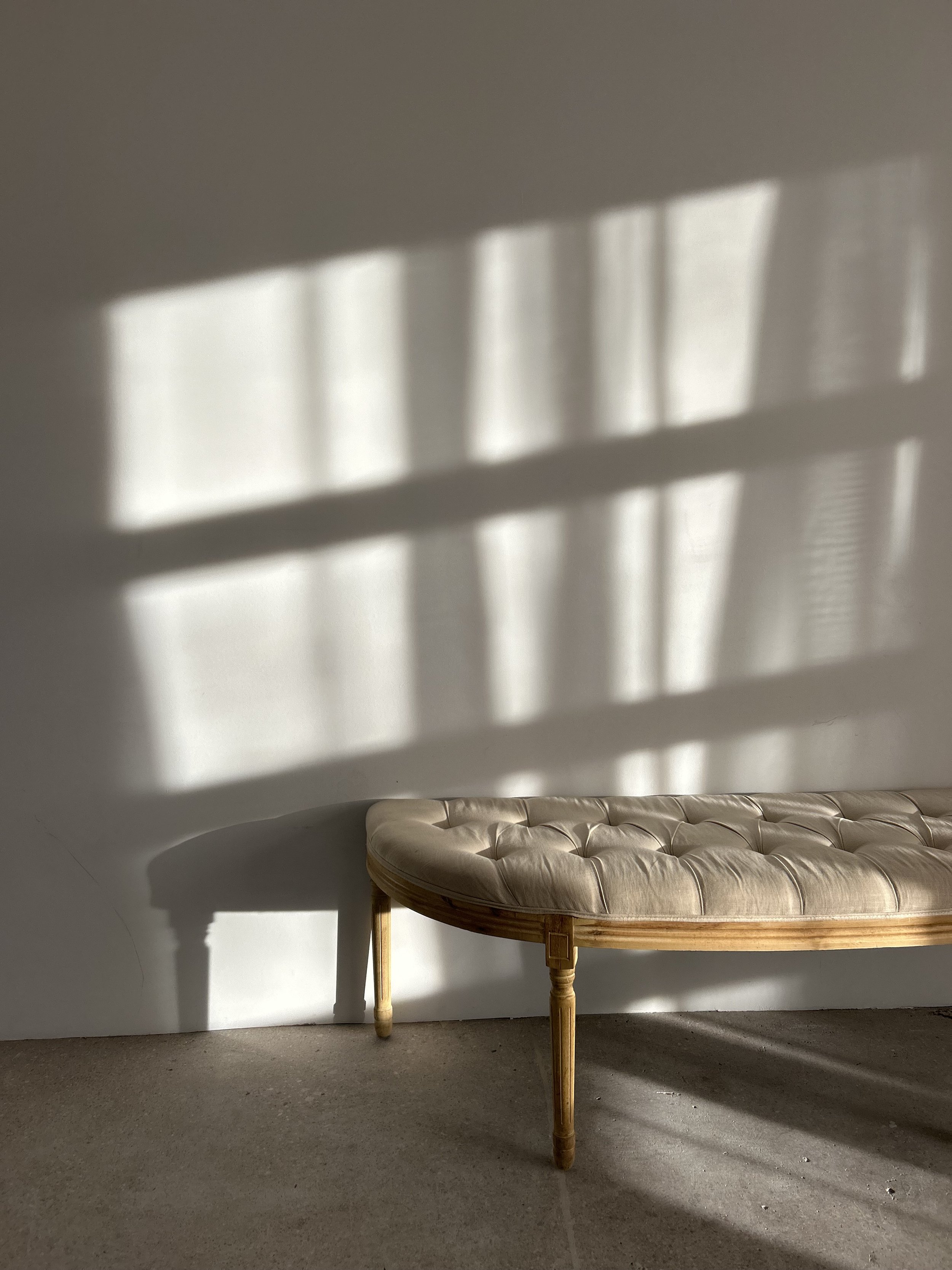 Sunlight creates shadow patterns on a plain wall, with a vintage beige tufted bench with wooden legs positioned in front of the wall.