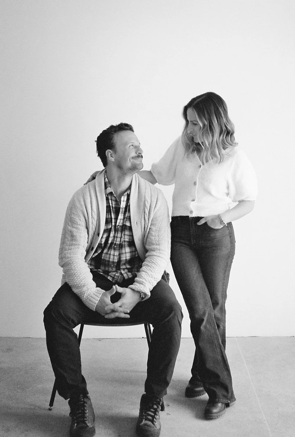 A man sitting on a chair looking up at a woman standing beside him, smiling. The woman is touching the man's shoulder, both in casual clothing, against a blank wall.