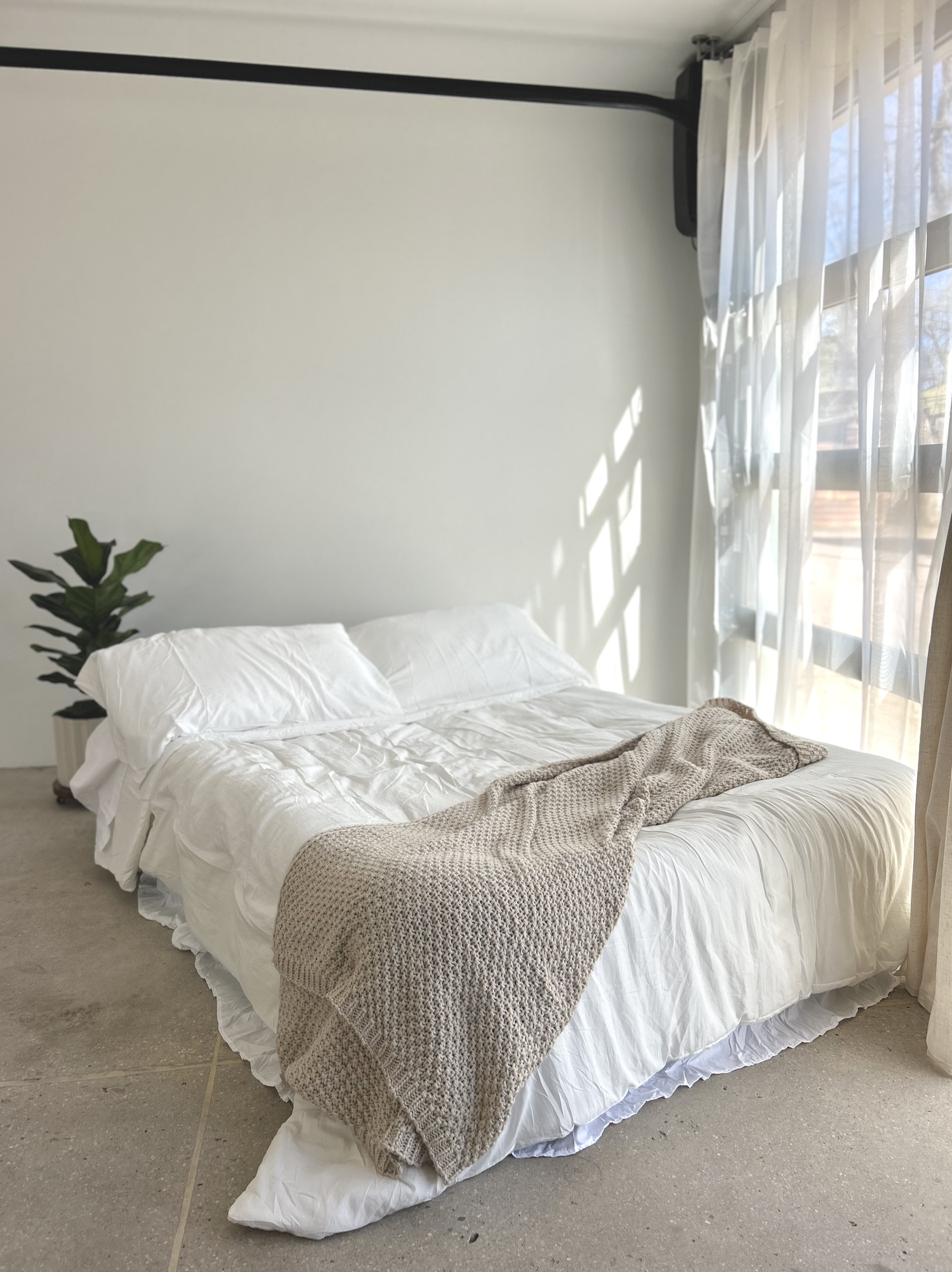 A neatly made bed with white bedding and pillows, a textured beige blanket draped over the bed, and a potted plant in the corner near a window with sheer curtains letting in natural light.