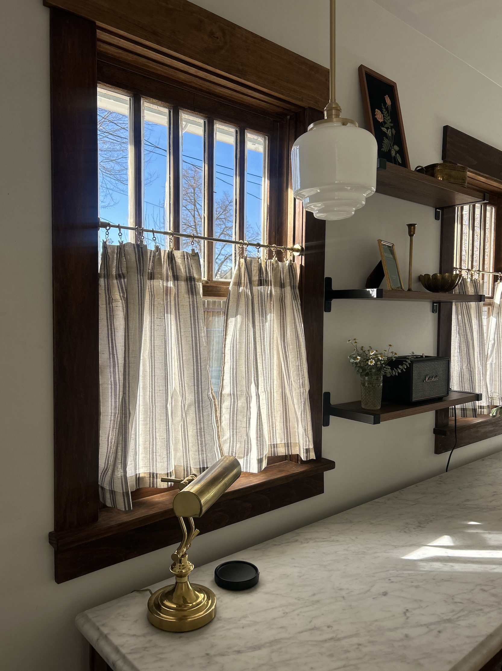 View of a window with wooden frame and striped curtains, a white pendant light hanging from the ceiling, a marble countertop with a brass desk lamp and a camera lens cap, and wall shelves with small decorative items and picture frames.