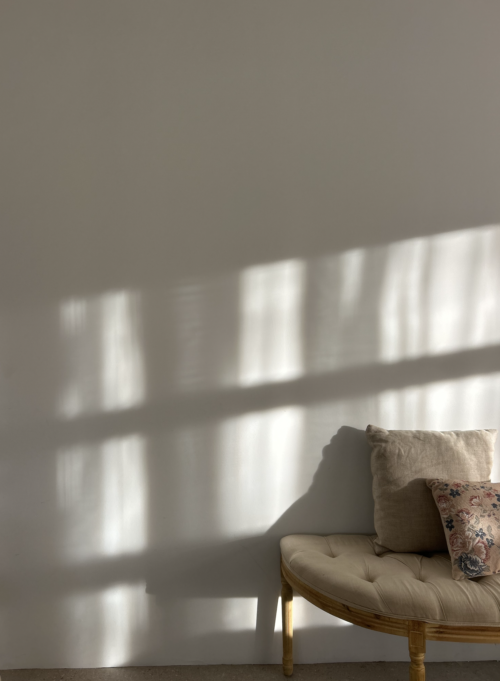 Sunlight casting shadows of window blinds on a plain wall with a beige cushioned chair and pillows at the bottom right.