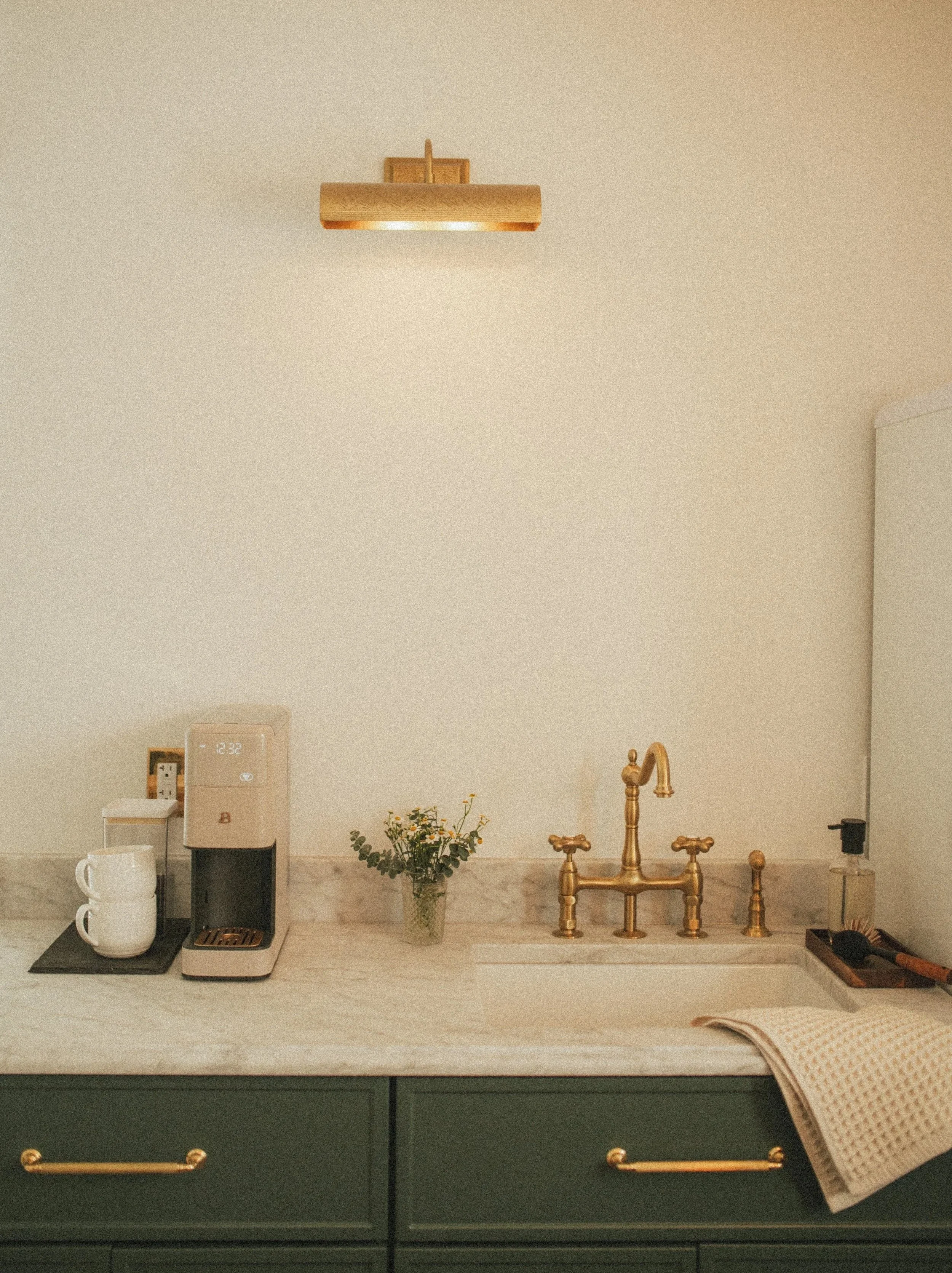 A kitchen countertop with a coffee machine, two white mugs, a small vase with flowers, a gold-colored faucet and sink, a soap dispenser, and a dish towel.
