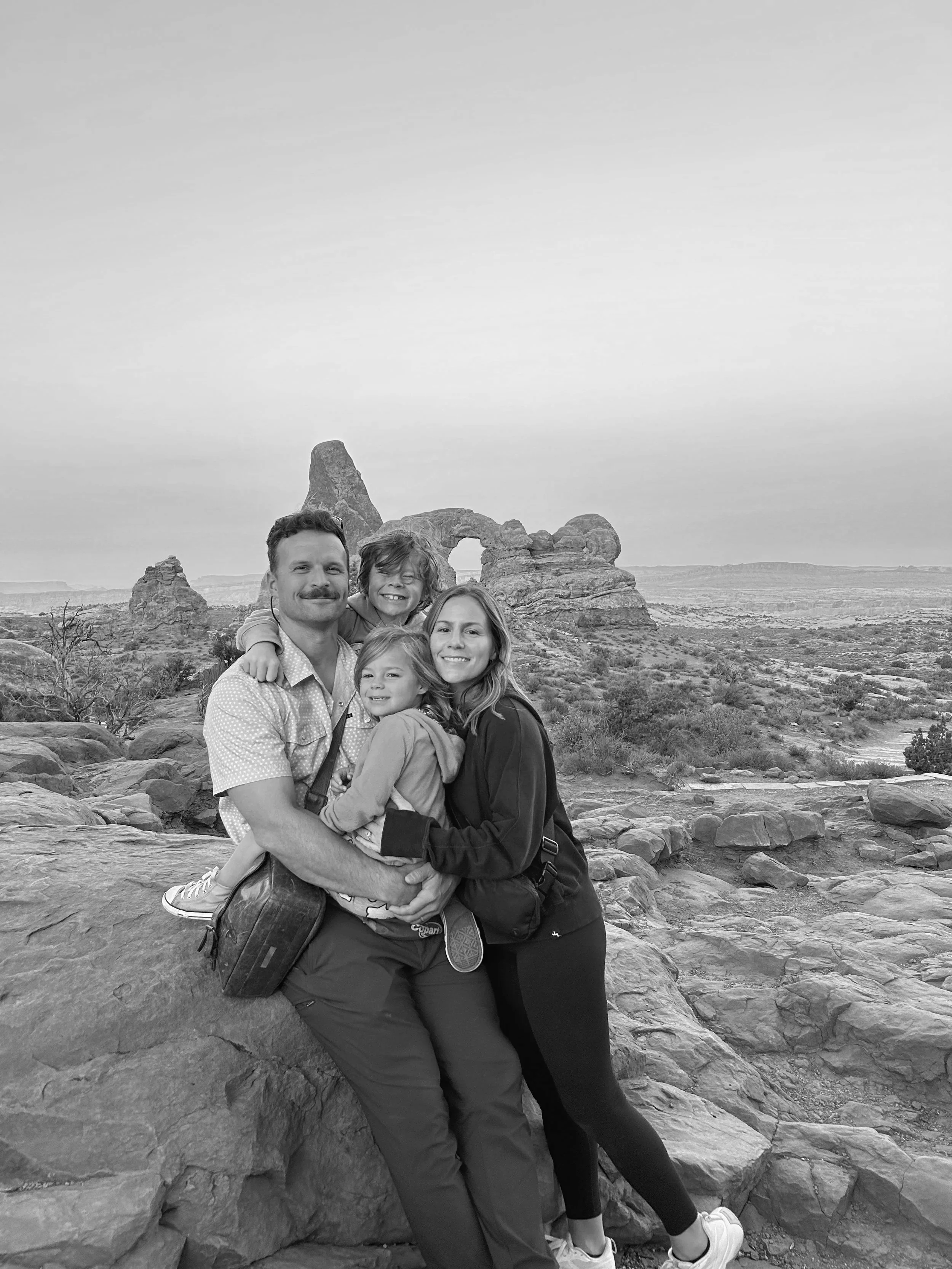 Family of four posing on rocky terrain with a natural rock formation and arch in the background, likely in a desert landscape.