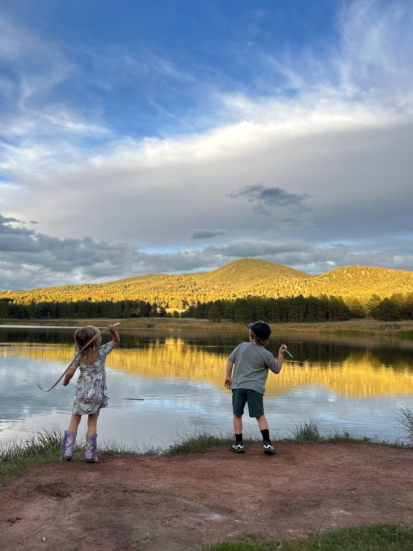 Two children, a girl and a boy, catch frogs by a lake at sunset with mountains and a partly cloudy sky in the background.