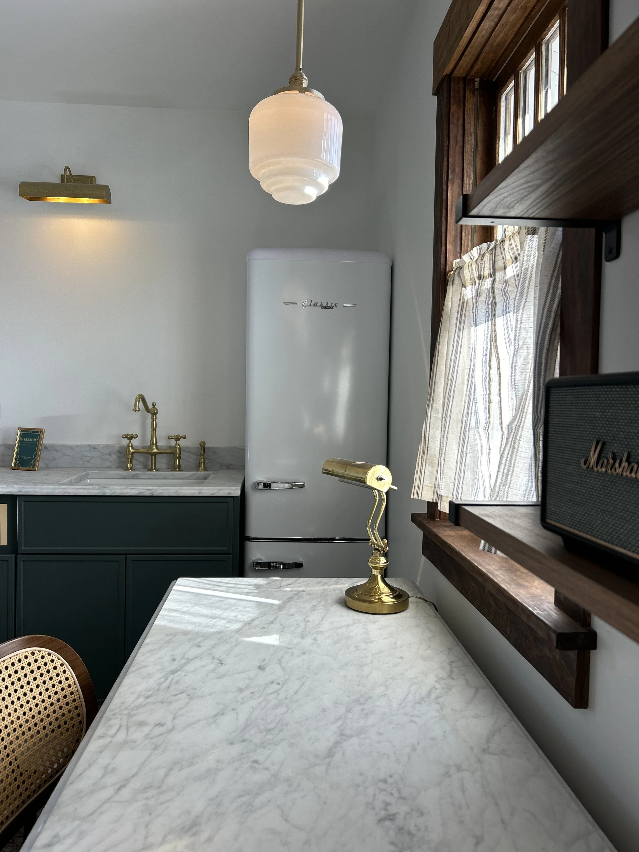A vintage-style kitchen with a marble countertop, brass fixtures, a white retro refrigerator, a brass desk lamp, dark green cabinetry, a marble sink area, a window with striped curtains, and a black Marshall speaker.