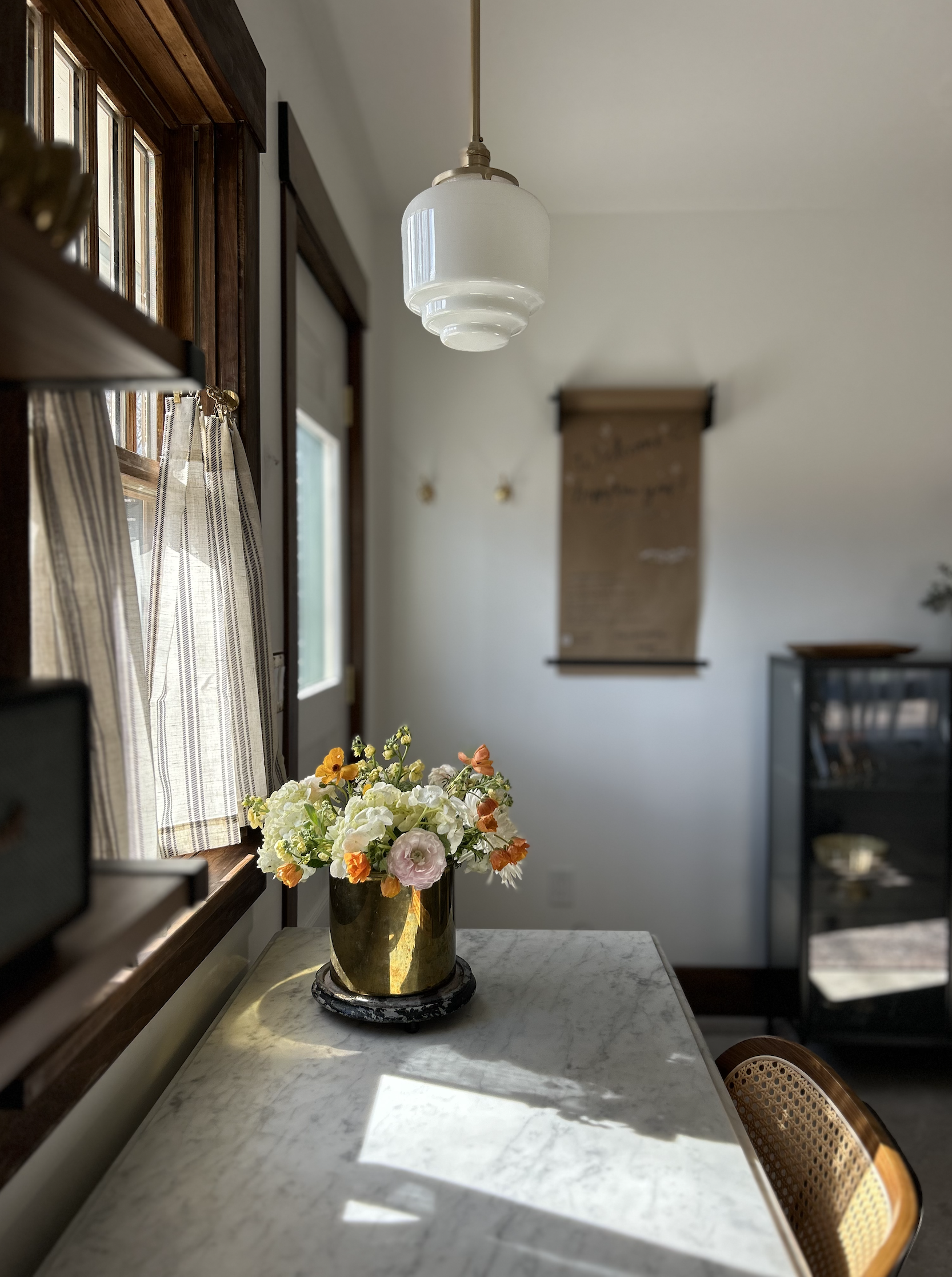 A marble-topped dining table with a gold flower vase filled with white, pink, and yellow flowers, near a window with striped curtains. A white hanging light fixture is above the table, and in the background, there is a wall with a scroll and a black 