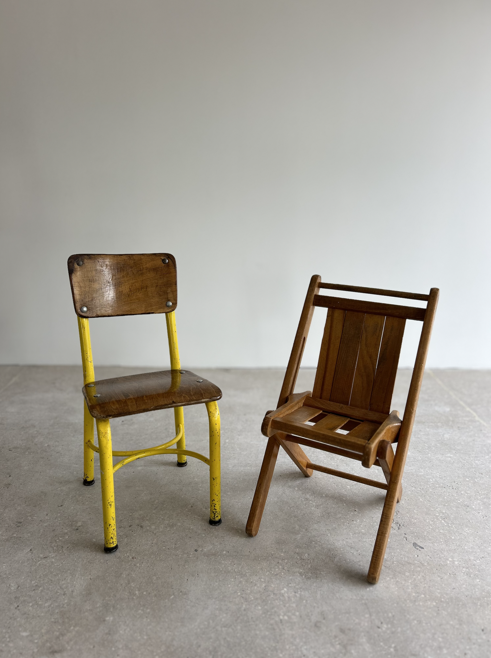 Two chairs, one with yellow metal frame and wooden seat and backrest, the other wooden with slatted seat and backrest, positioned on a concrete floor against a plain wall.