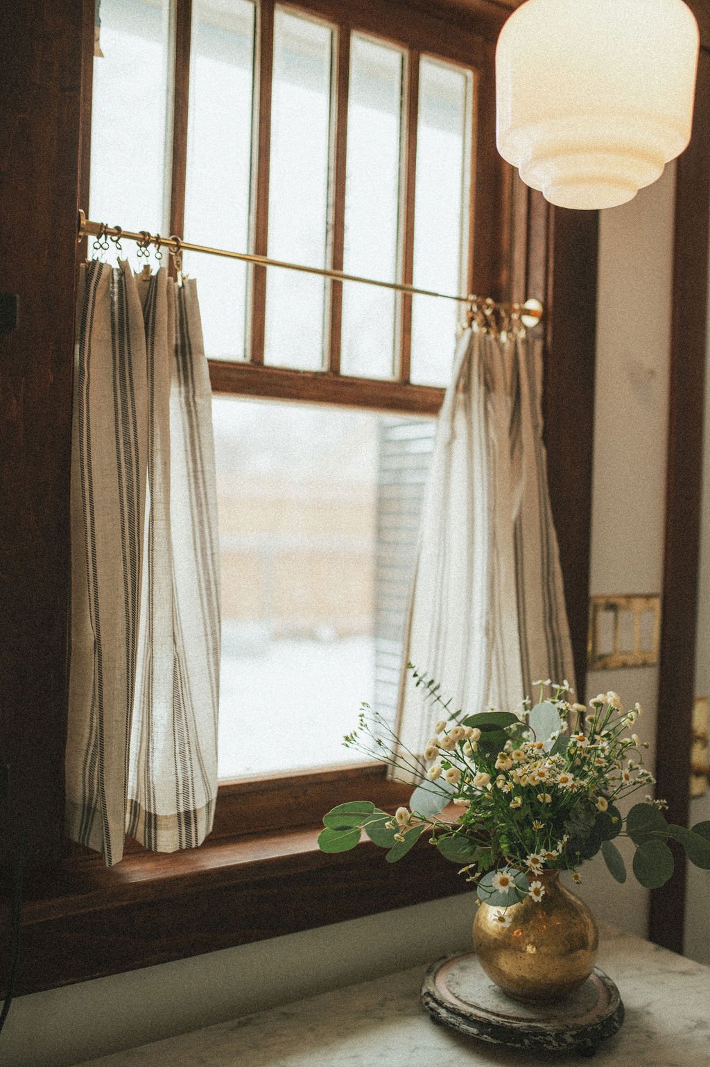 A window with wooden framing and striped curtains, with a bouquet of flowers in a gold vase on a windowsill.