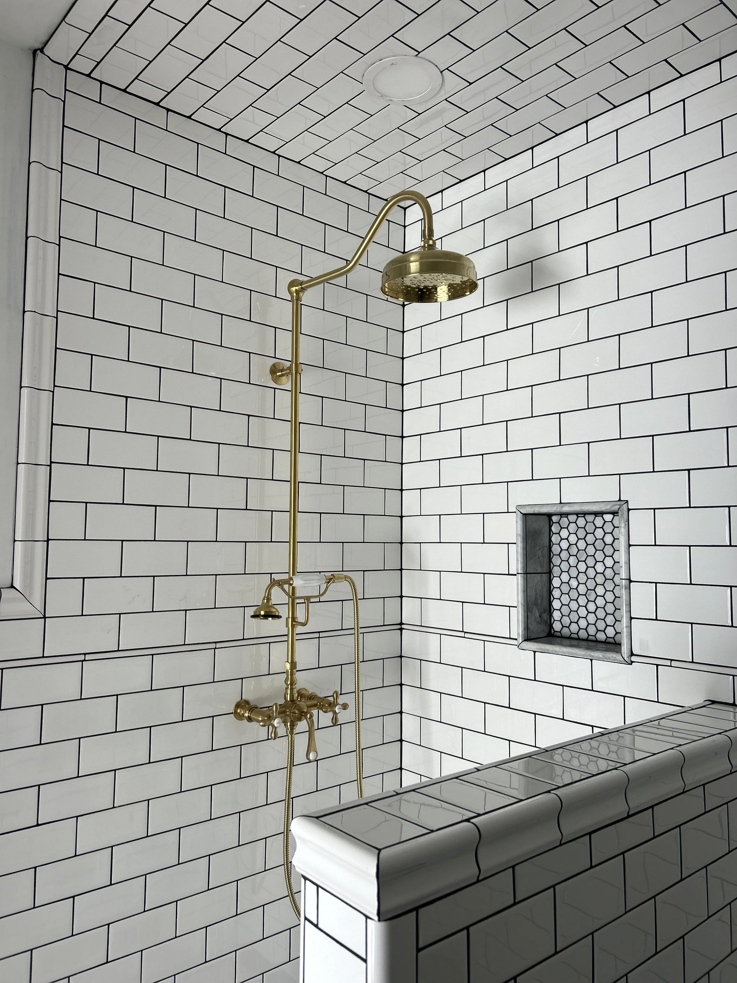 A shower area with white subway tile walls, a brass shower fixture, and a built-in tiled shelf.