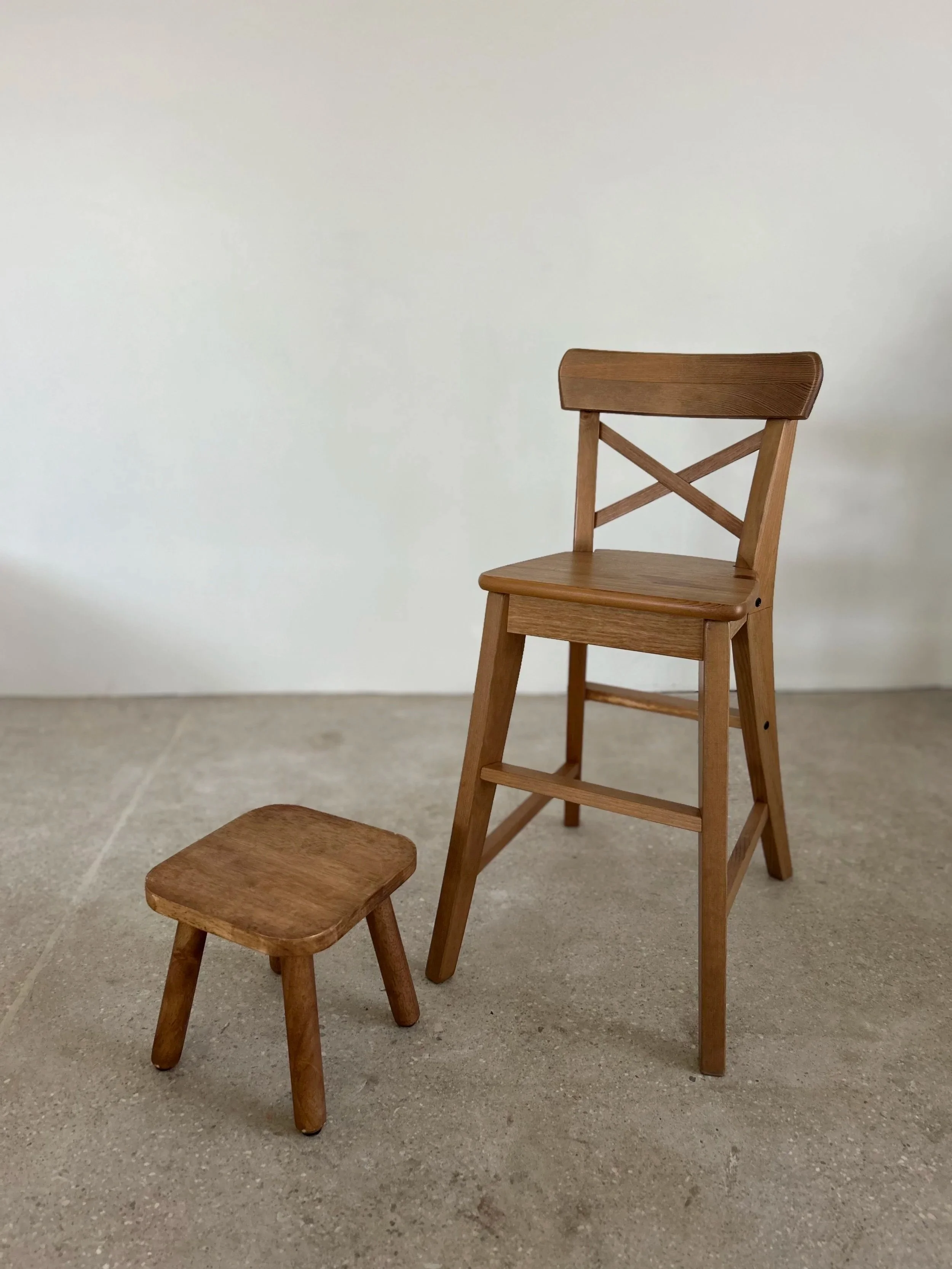 Two wooden chairs, one tall with a cross-back design and one low stool, are placed on a concrete floor against a plain white wall.