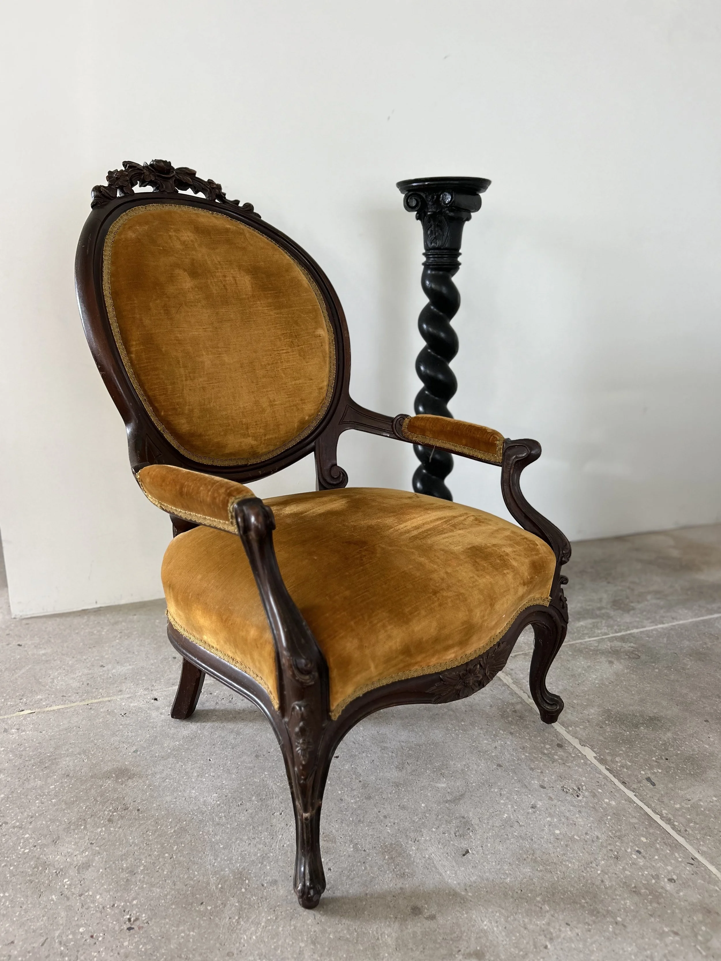 An antique upholstered armchair with a wooden frame and velvet fabric, positioned next to a tall black twisted pedestal table against a plain white wall.