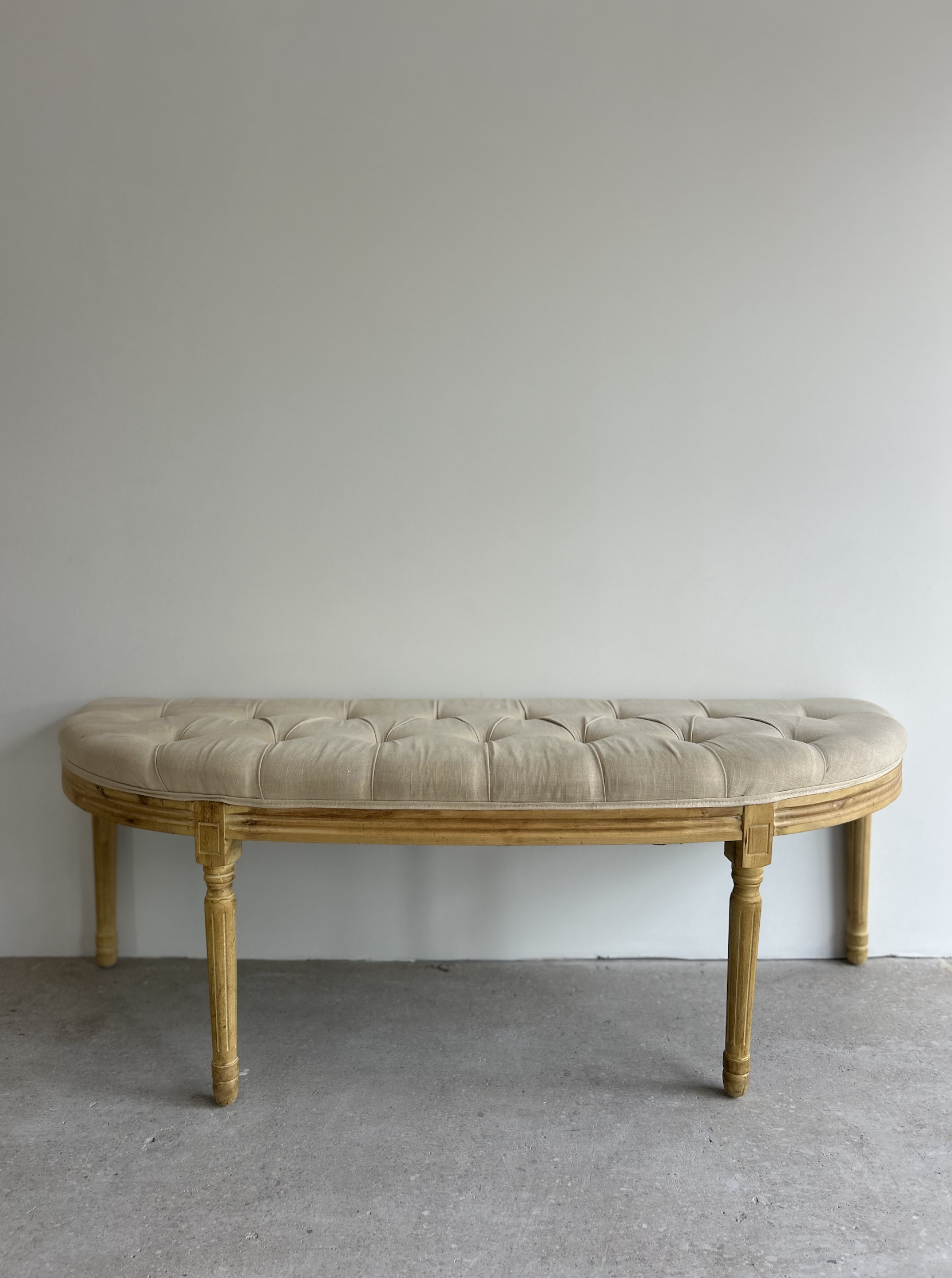 A vintage wooden bench with a cushioned, tufted beige seat against a plain white wall on a concrete floor.