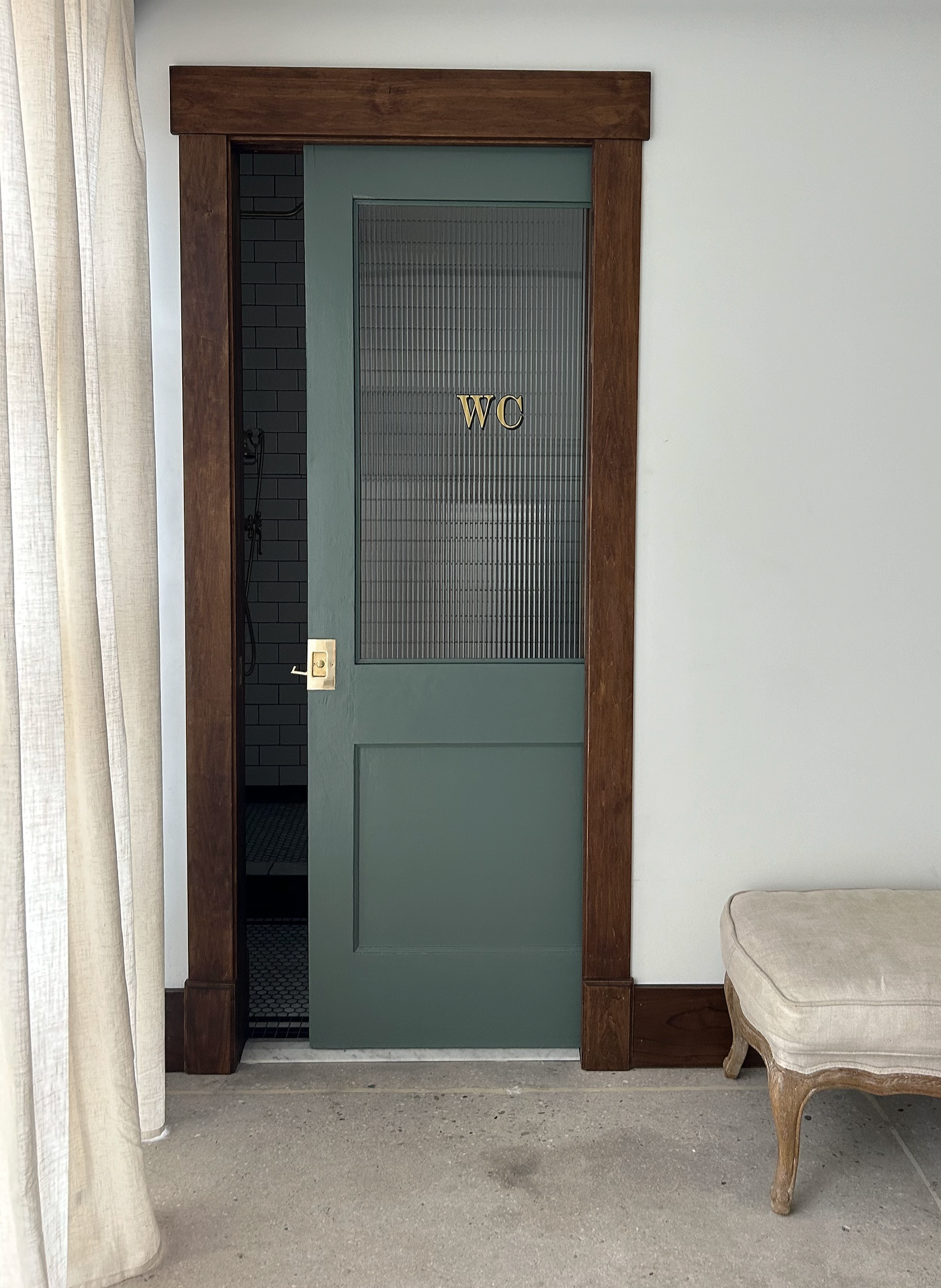 Green door with glass panel labeled 'WC' and a wooden frame, partly open revealing a shower with black tiles inside, adjacent to a beige cushioned bench with wooden legs, and a cream-colored curtain on the left side.