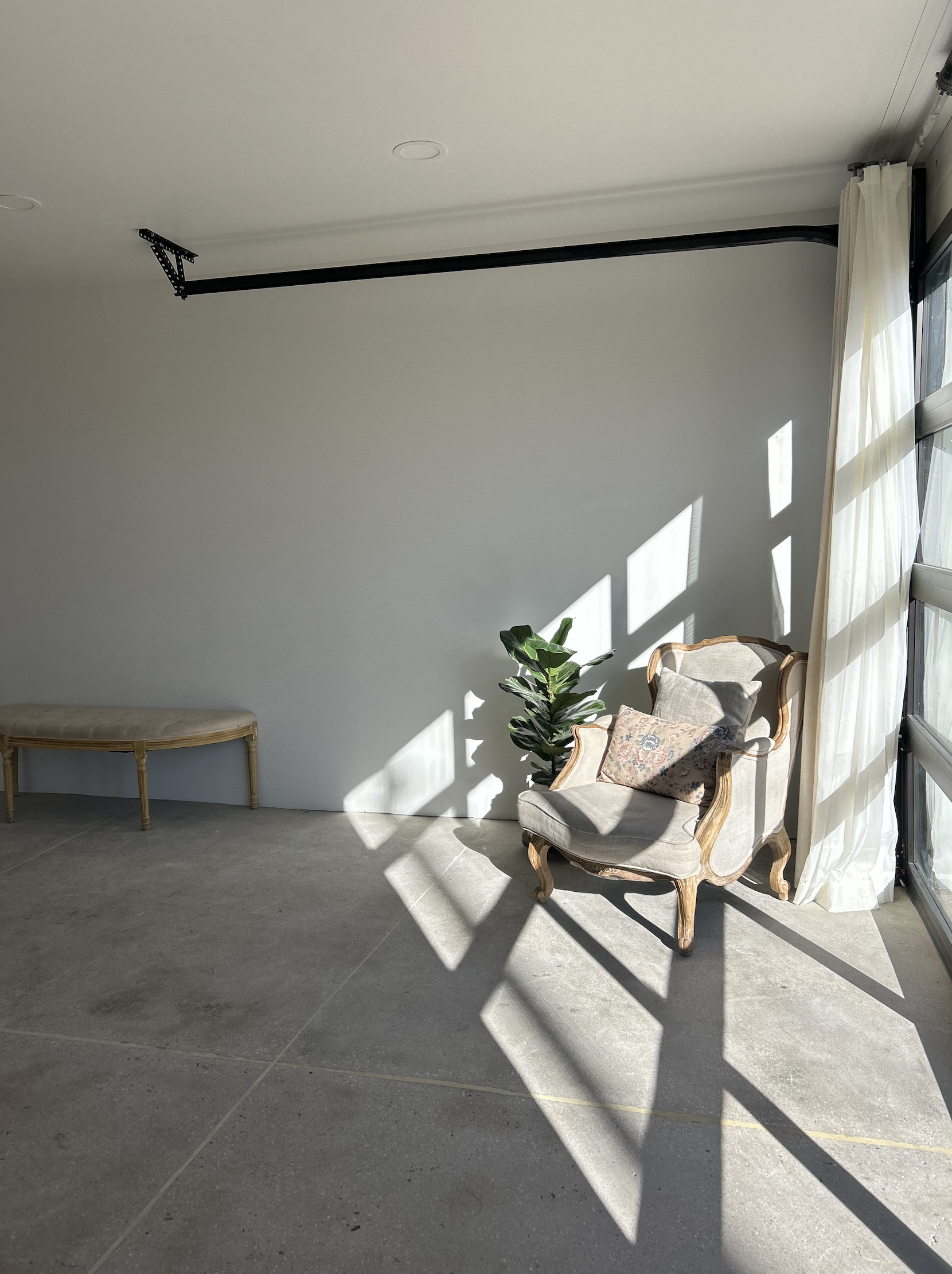 Sunlight casting shadows in a minimalist room with a vintage armchair, a small bench, and a potted plant near large windows with white curtains.