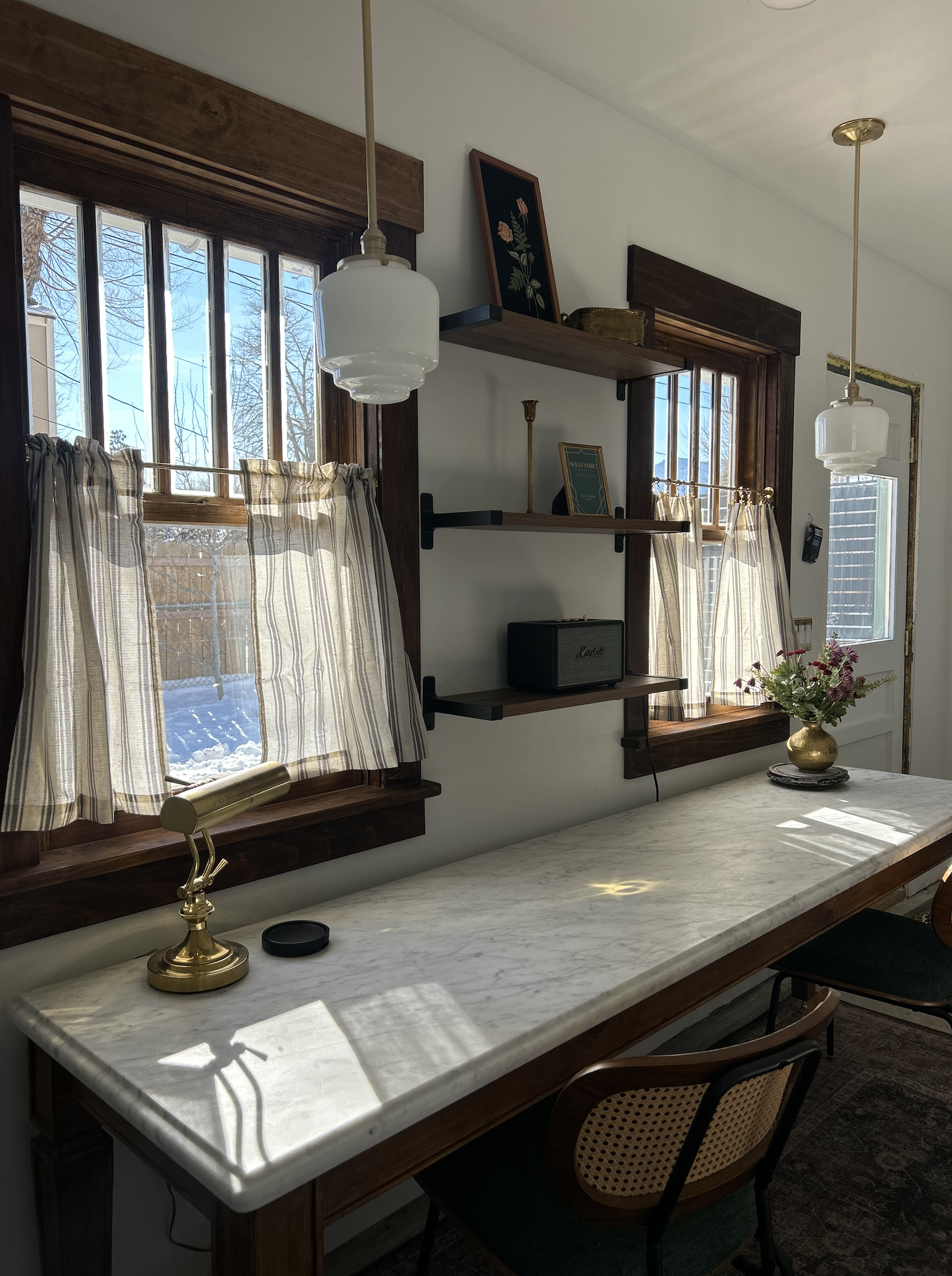 A cozy interior with a marble-topped wooden table, two windows with striped curtains, a brass desk lamp, a vase with flowers, wall-mounted black shelves, and framed decor.