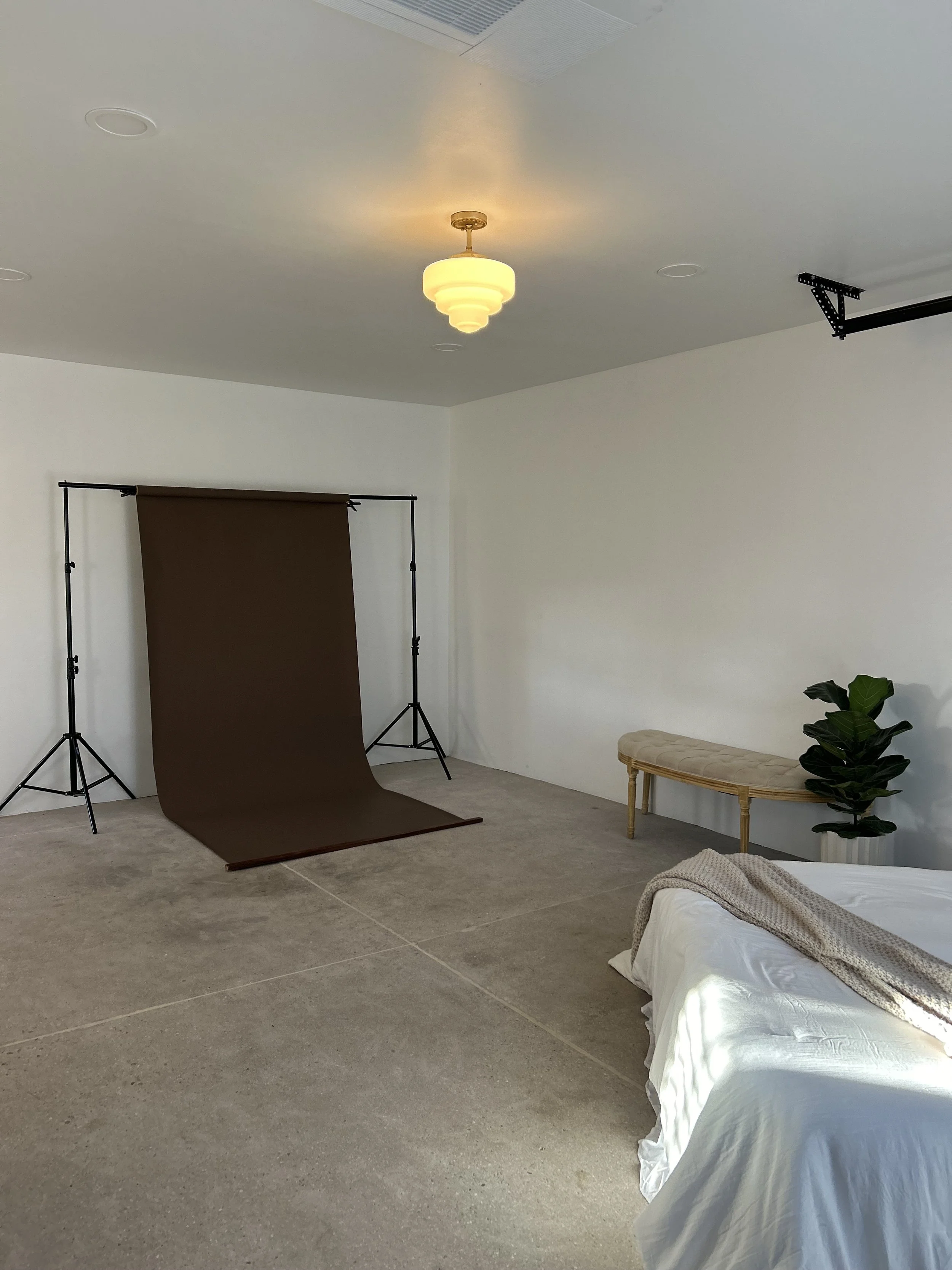 A room with a brown photography backdrop stand, a small bench, a plant, a bed with white bedding and a beige throw, and a ceiling light fixture.