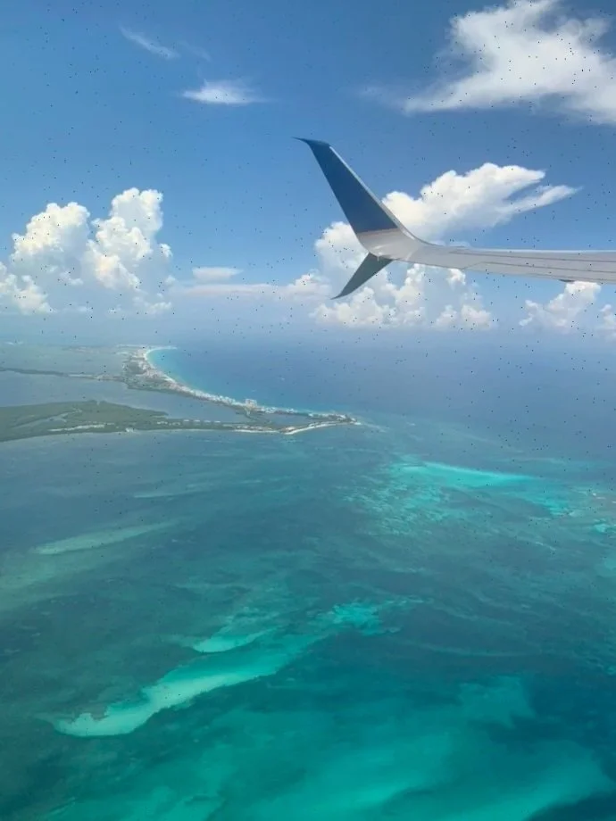 View from an airplane window showing a blue ocean, coral reefs, and a coastline with some land, under a partly cloudy sky.