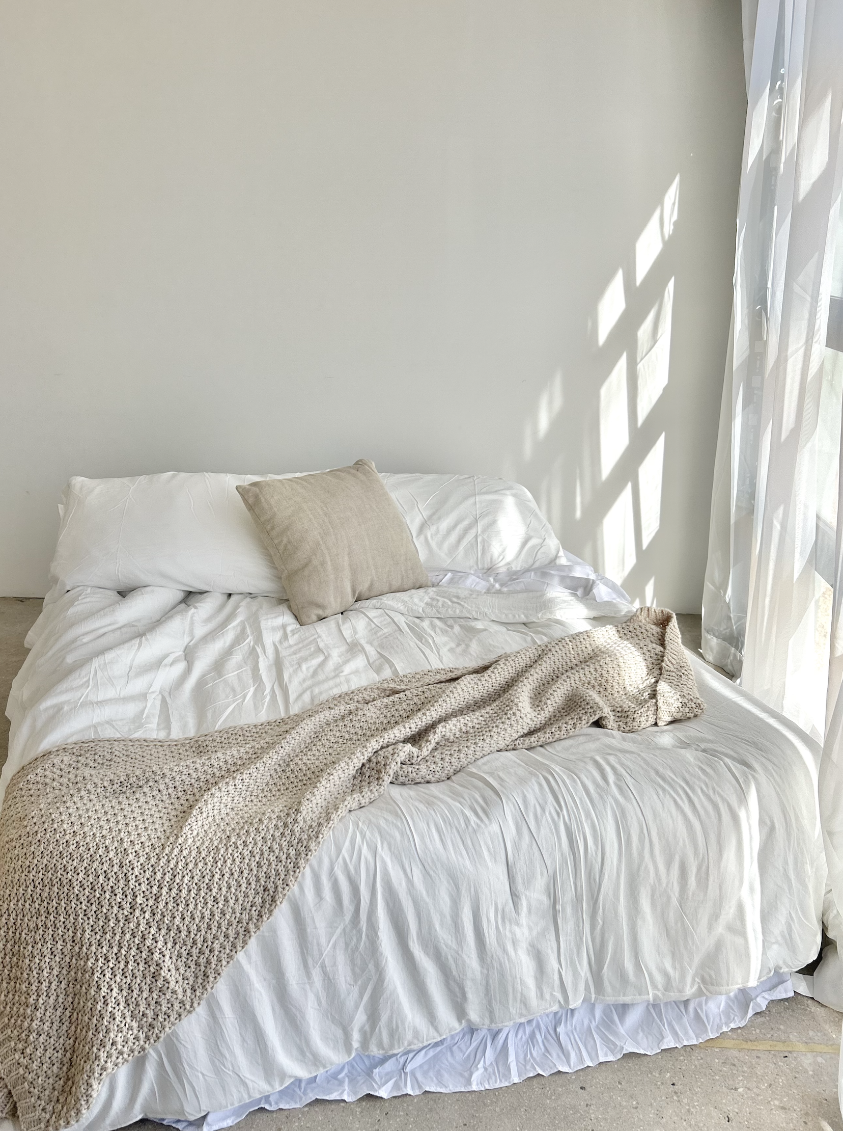 A neatly made bed with white bedding, a beige pillow, a textured beige blanket, and diffused sunlight coming through a sheer curtain on the right.