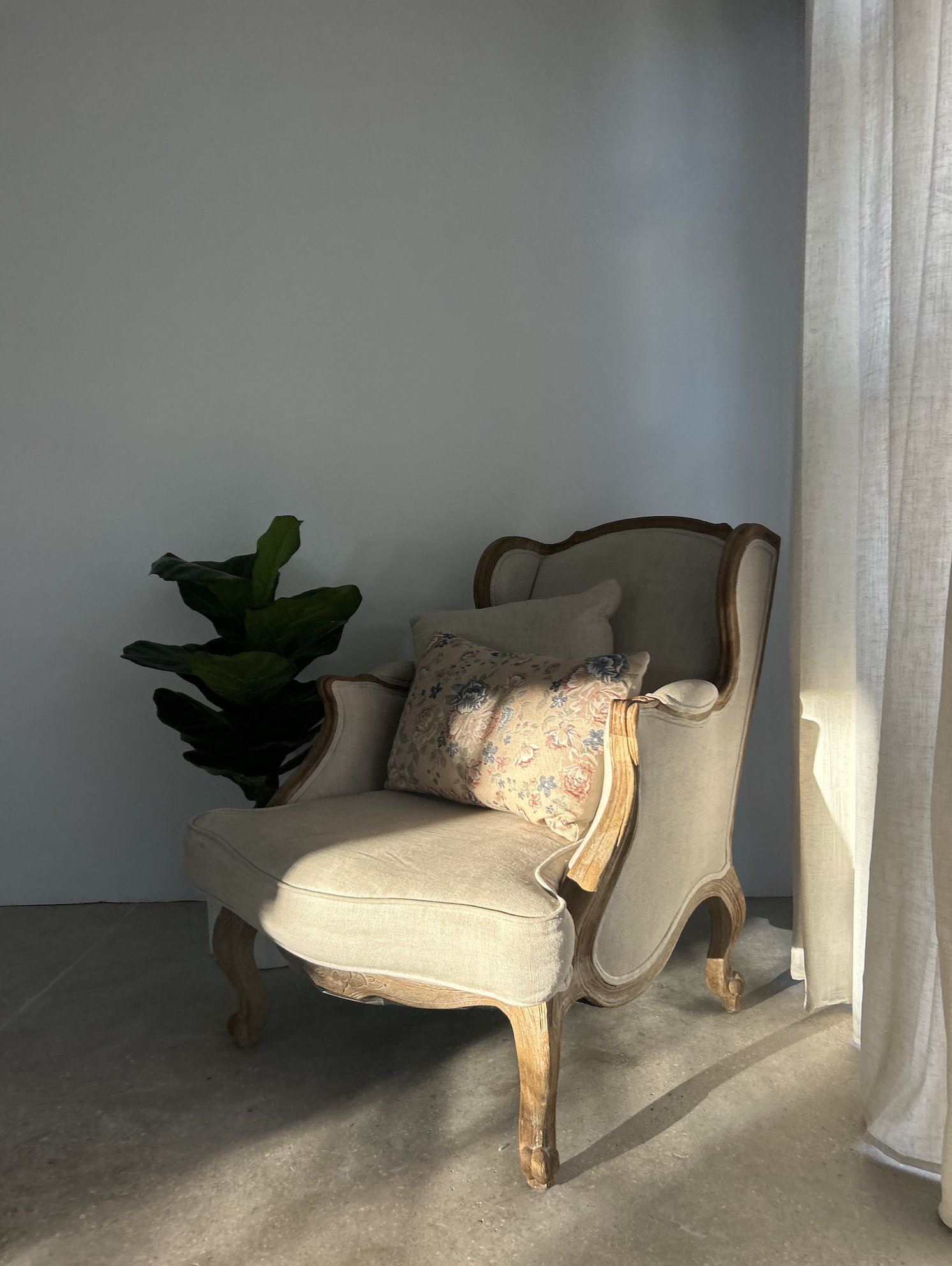An antique beige upholstered armchair with wooden frame and carved legs, placed next to a green potted plant and cream-colored curtains with sunlight streaming in.