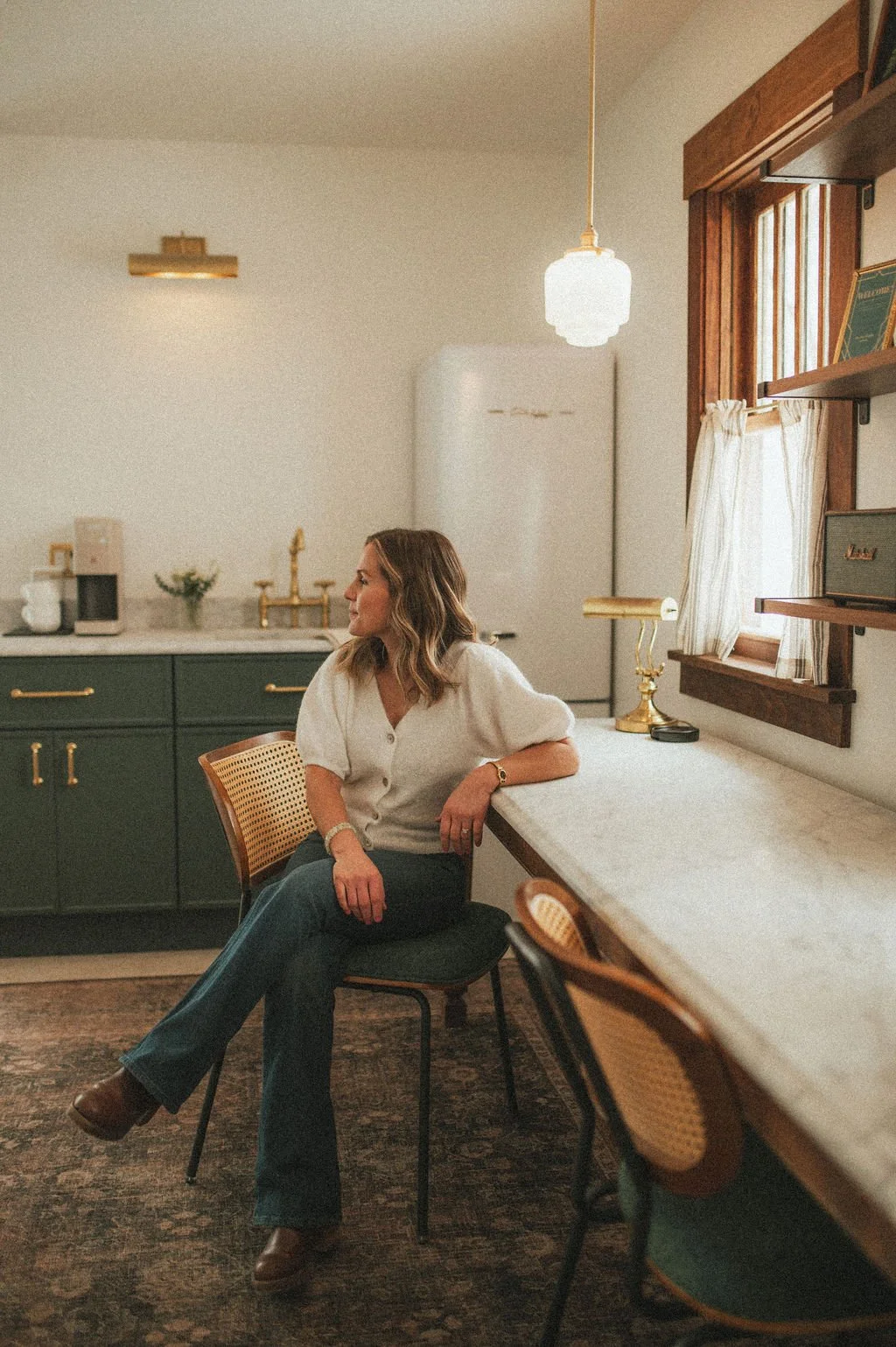 A woman sitting on a chair in a cozy, well-decorated kitchen with vintage and modern elements, looking out the window with a relaxed expression.