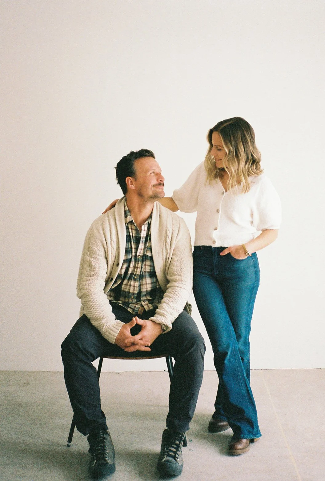 A man and a woman in casual clothing interacting, with the man sitting on a chair and the woman leaning on his shoulder, both smiling at each other against a plain white background.