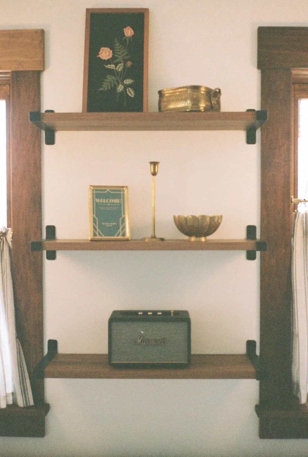 Wooden wall shelf with three levels holding decorative items, framed art, and a small amplifier. The wall has two windows with curtains on each side.