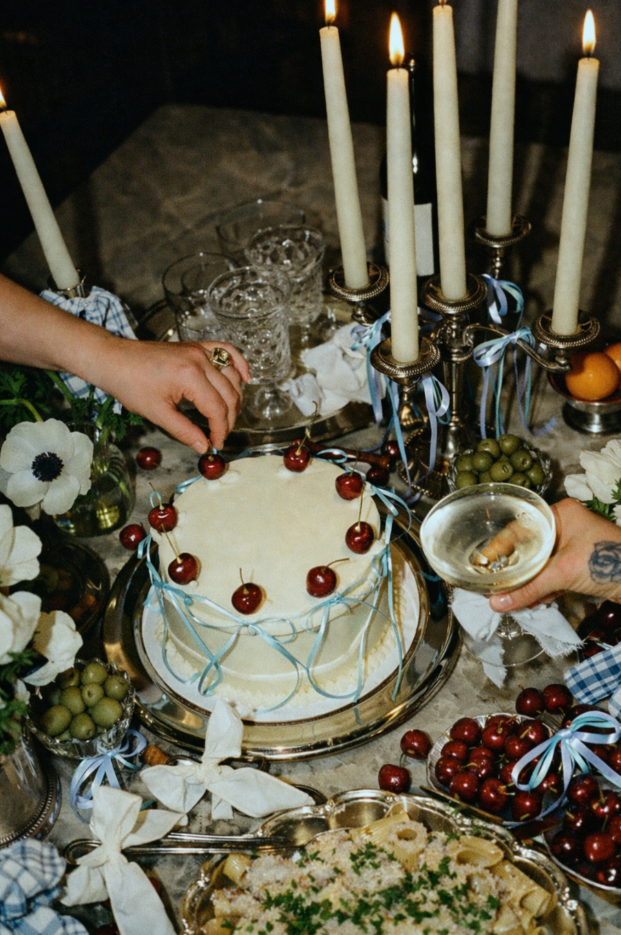 A table decorated for a celebration with a white frosted cake topped with cherries, surrounded by glasses, flowers, grapes, and candles.