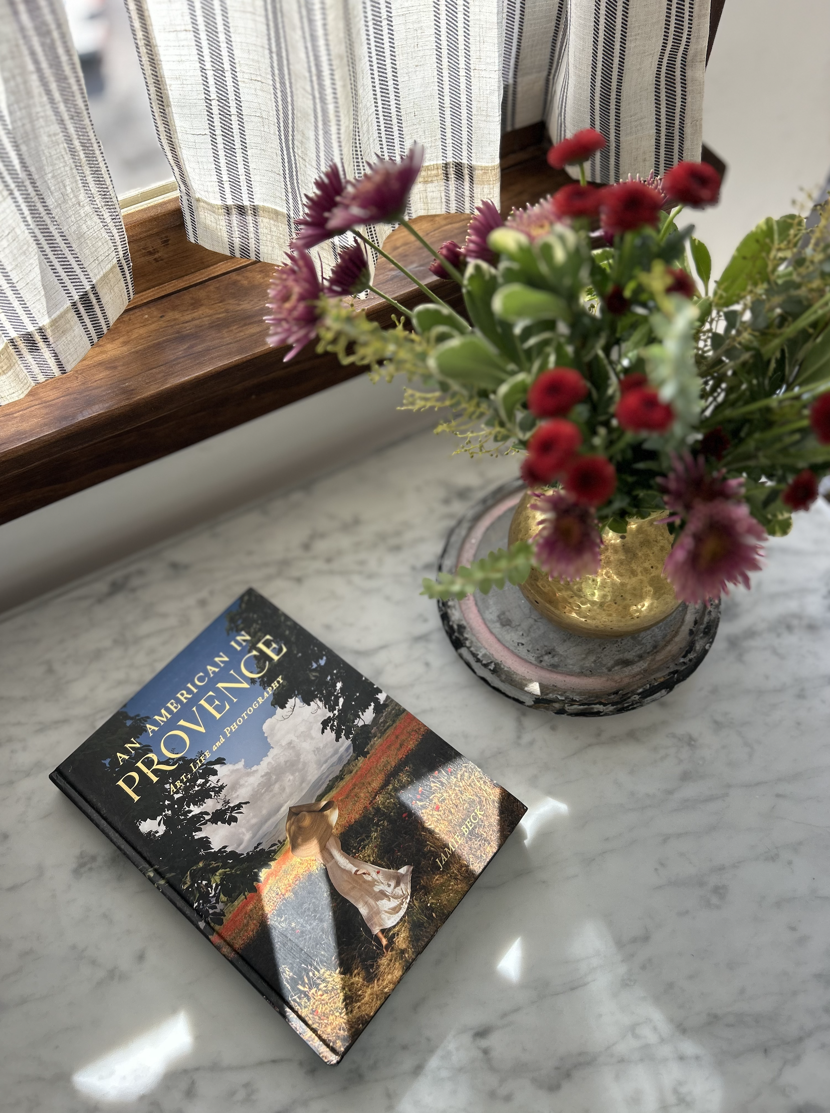 A book titled "An American in Provence" lies on a white marble table next to a vase of pink and red flowers. The table is near a window with striped curtains, allowing natural light to illuminate the scene.