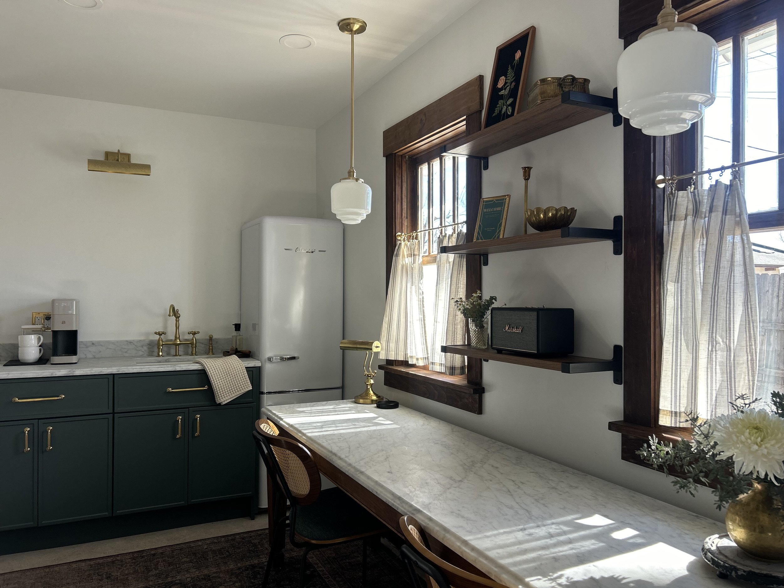 A bright kitchen with green cabinets, a marble countertop, gold fixtures, white curtains, wooden shelves with decorative items, and natural light coming through windows.