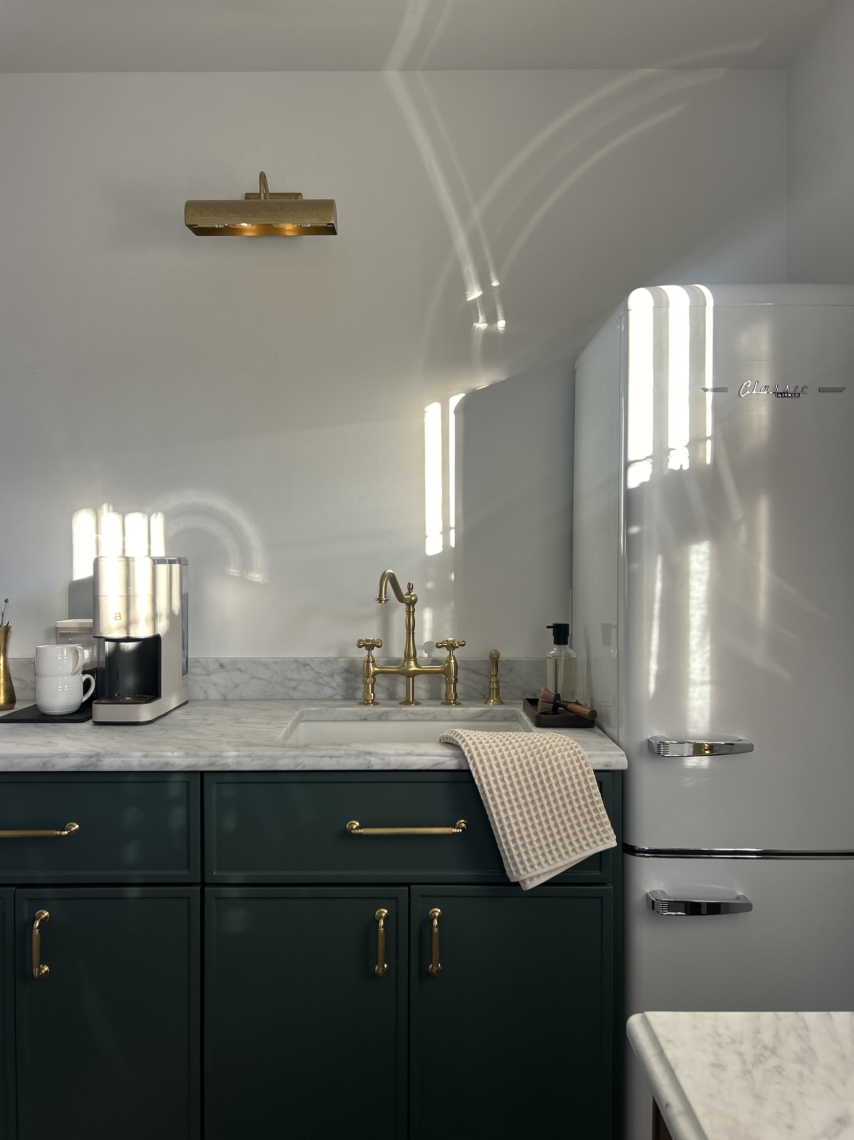 Kitchen with dark green cabinets, marble countertop, gold hardware, gold faucet, white marble backsplash, white vintage fridge, coffee maker, and cups, with sunlight casting shadows on the wall.