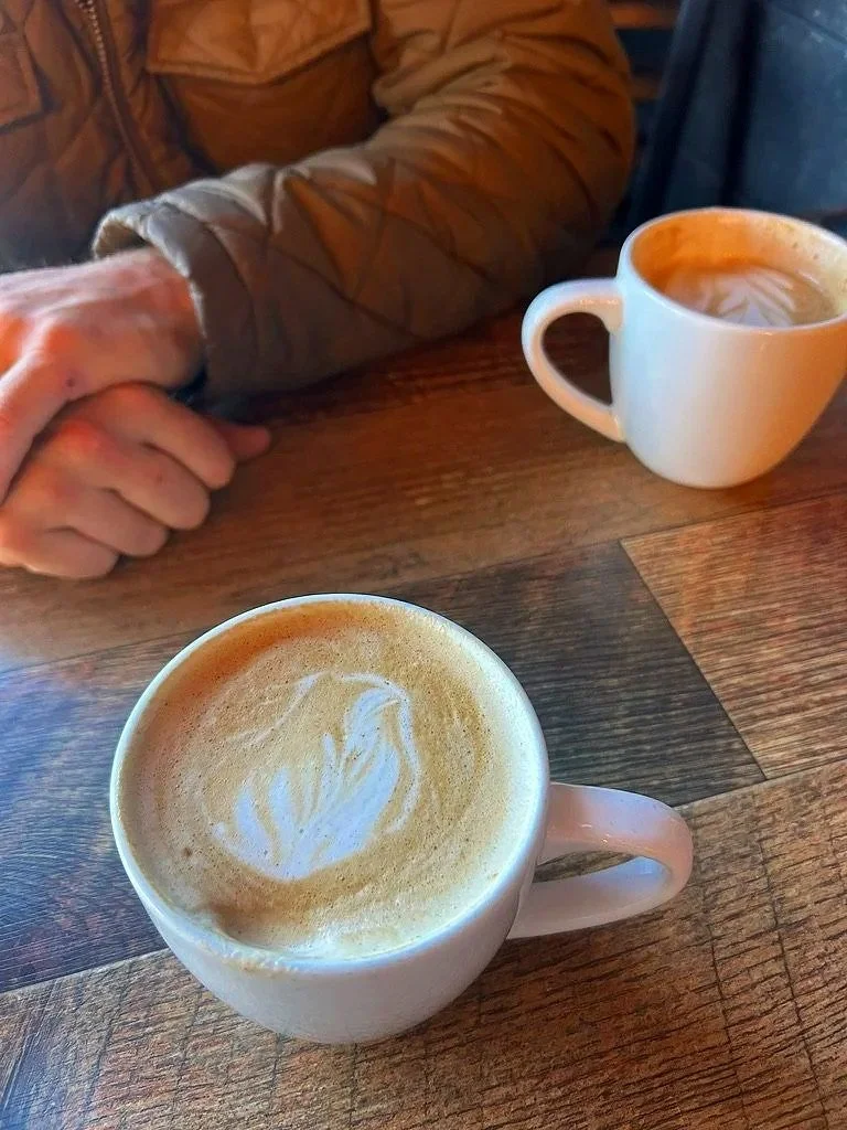 Two cups of coffee with latte art on a wooden table, with a person's arm and hand resting nearby.
