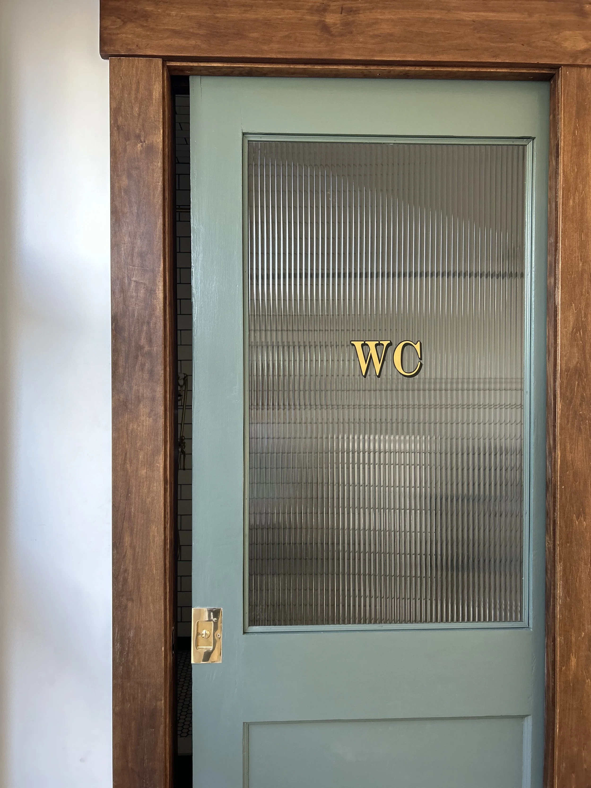 A light green door with a brass handle and glass panel that has the letters 'WC' in yellow, indicating a water closet or restroom. The door is surrounded by wooden trim.