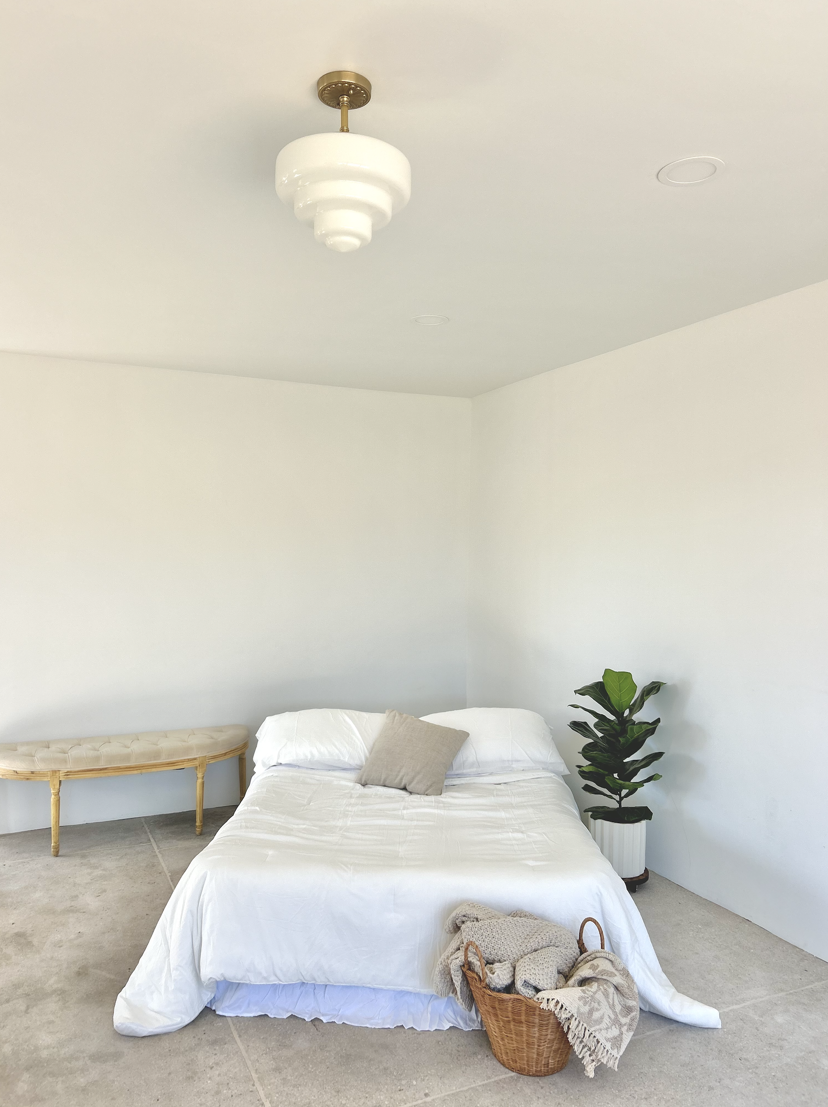 A minimalist bedroom with a white bed, a beige pillow, a wicker basket with blankets, a potted fiddle leaf fig plant, a white bench, and a ceiling light fixture.