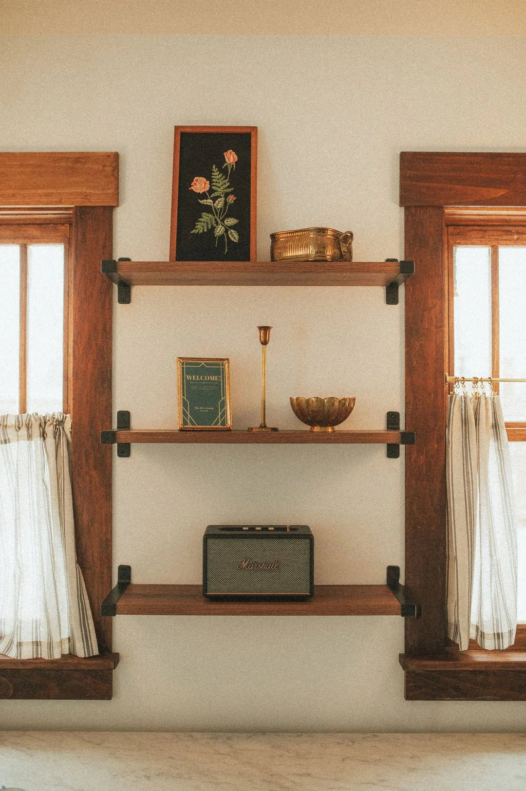 Wooden wall shelves with decorative items between two windows with plaid curtains.