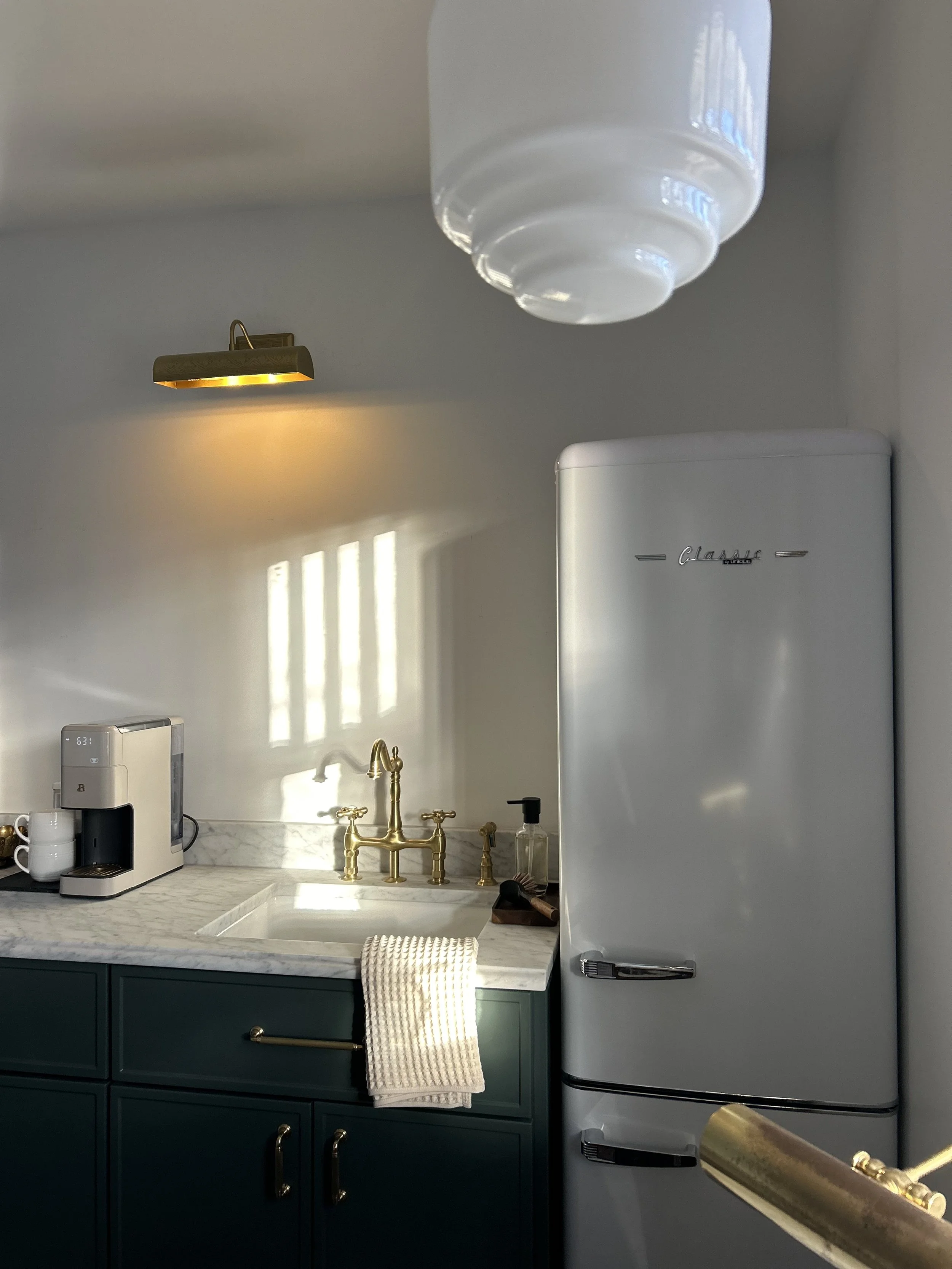 A modern kitchen corner with a white retro-style refrigerator, a marble countertop, a gold faucet, a coffee machine, a towel, a soap dispenser, and a wall-mounted light fixture.