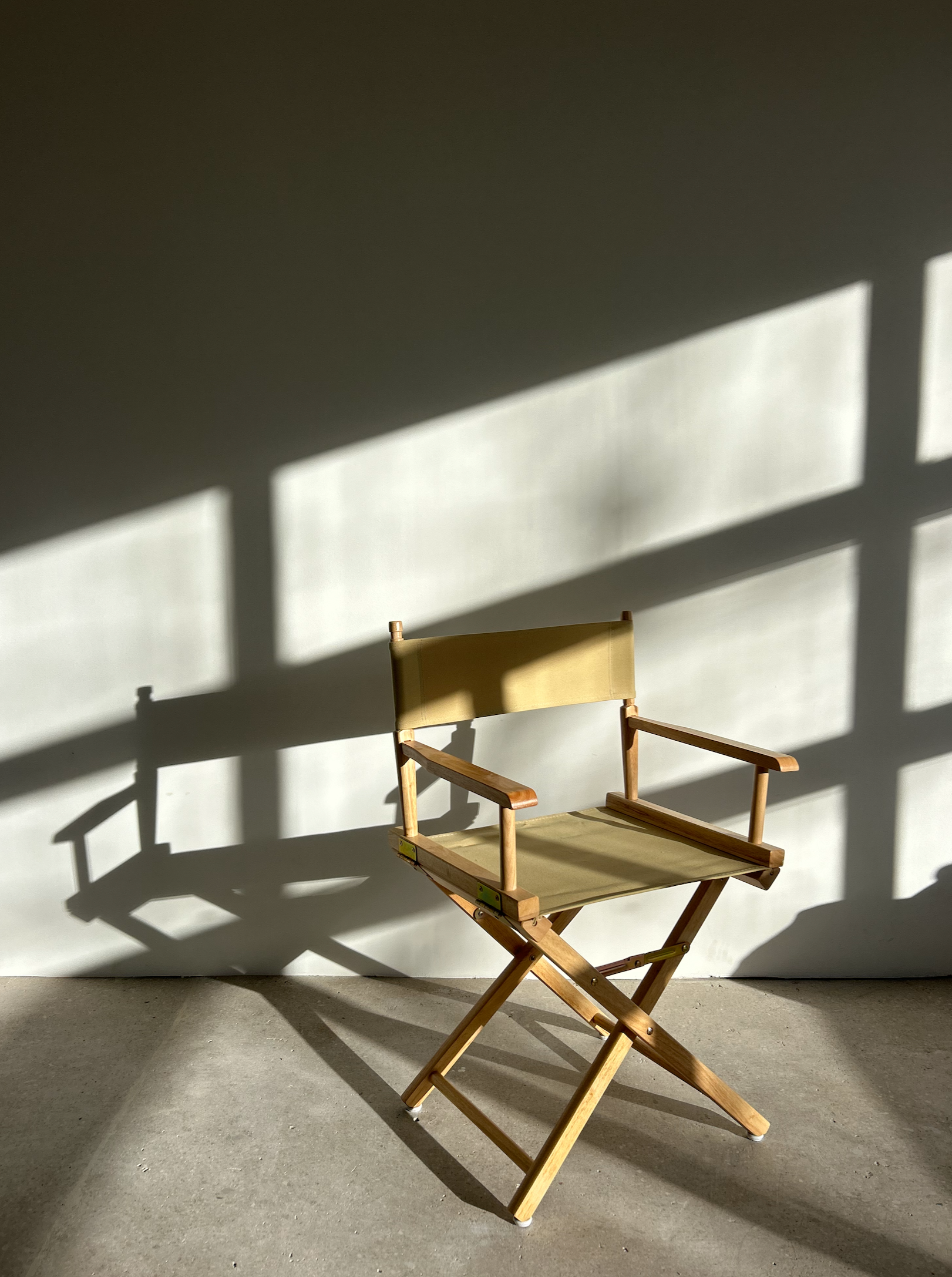Wooden director's chair with light beige fabric, casting a shadow on a white wall, illuminated by angled sunlight.