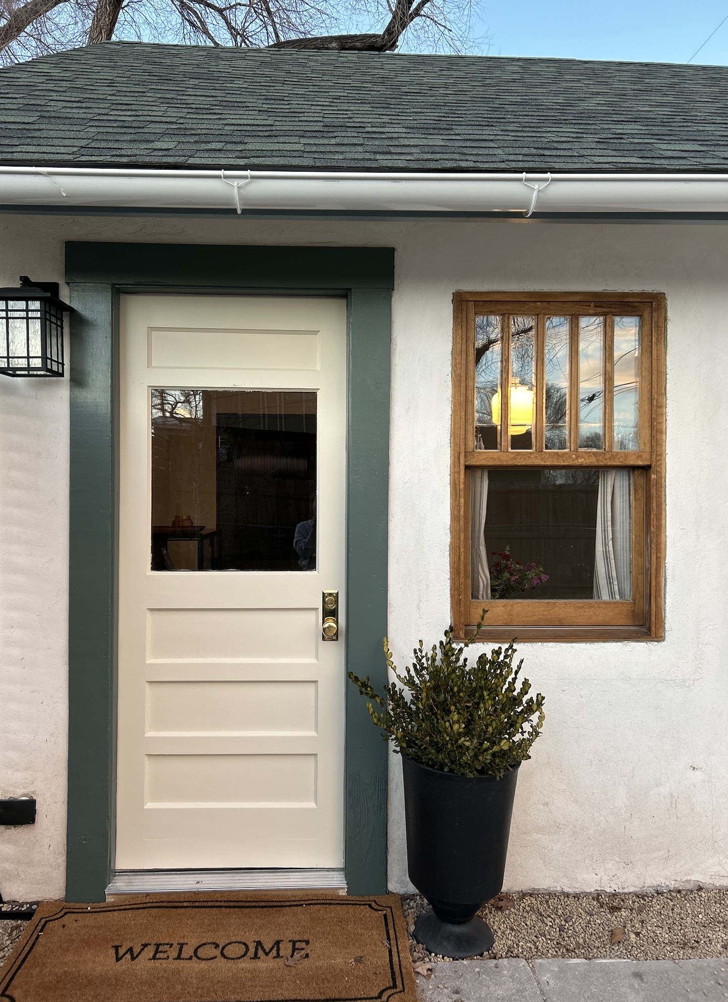 Front door of a house with a beige door, green trim, a window with a flowerpot inside, a black outdoor light, a potted plant, and a welcome mat.