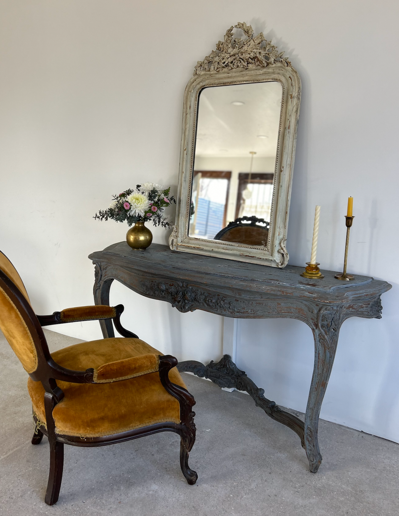 Vintage wooden vanity table with ornate carvings, topped with a large rustic mirror, a small flower vase, and two candlesticks, accompanied by an antique armchair with gold upholstery.